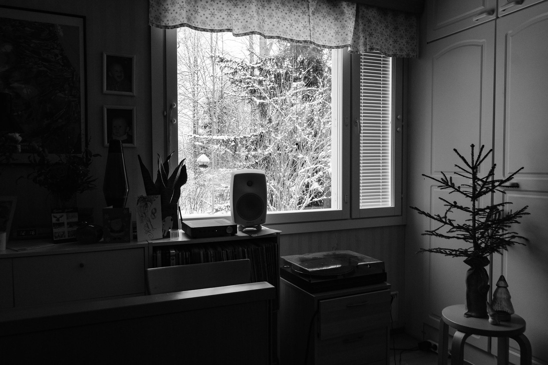 A black and white photo of a window view revealing a snowy landscape. Inside the room, there is a small Christmas tree on top of a stool, a turntable and a speaker, and various personal belongings on top of a sideboard.