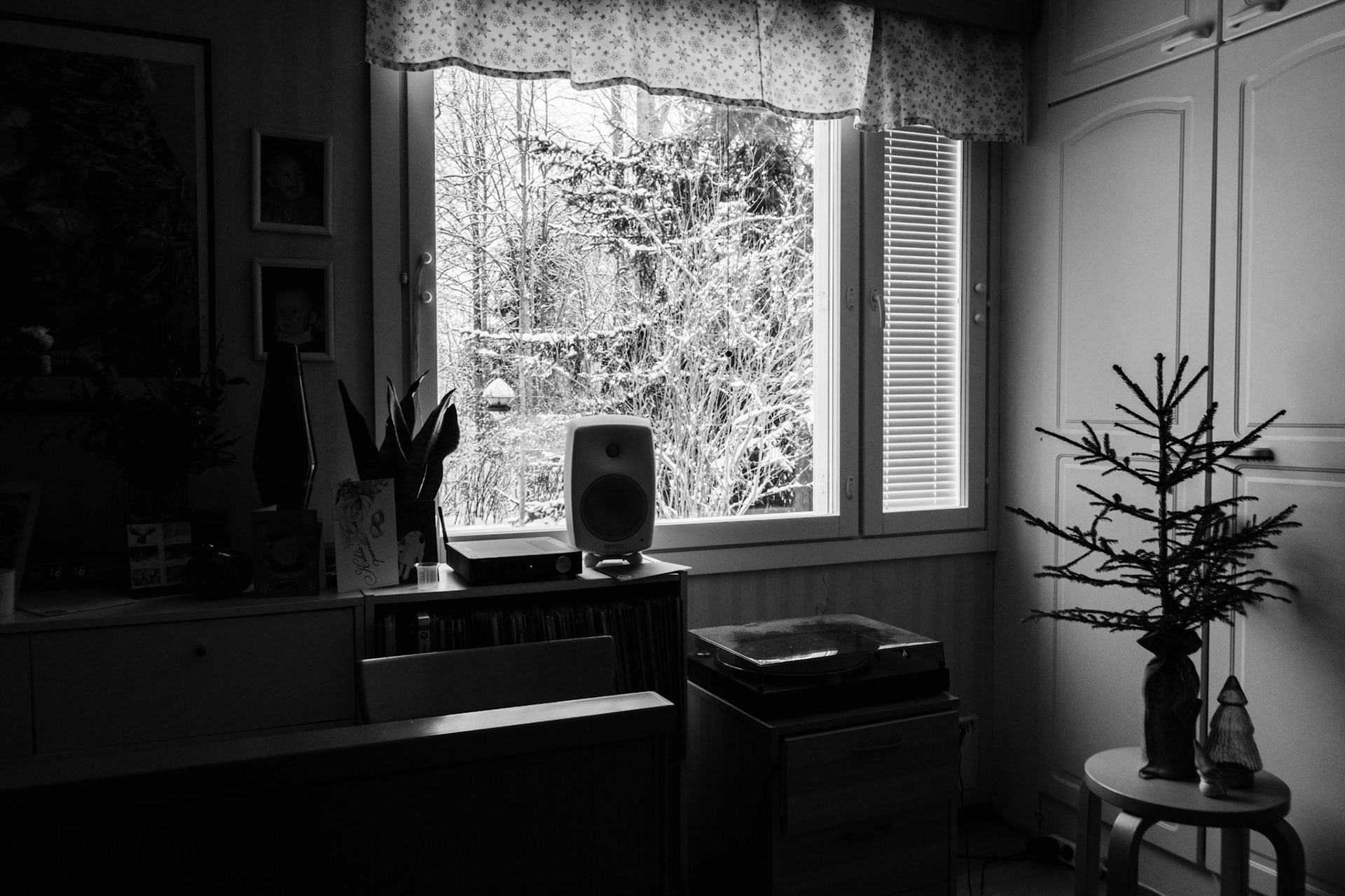 A black-and-white photo of a window view revealing a snowy landscape. Inside the room, there is a small Christmas tree on top of a stool, a turntable and a speaker, and various personal belongings on top of a sideboard.