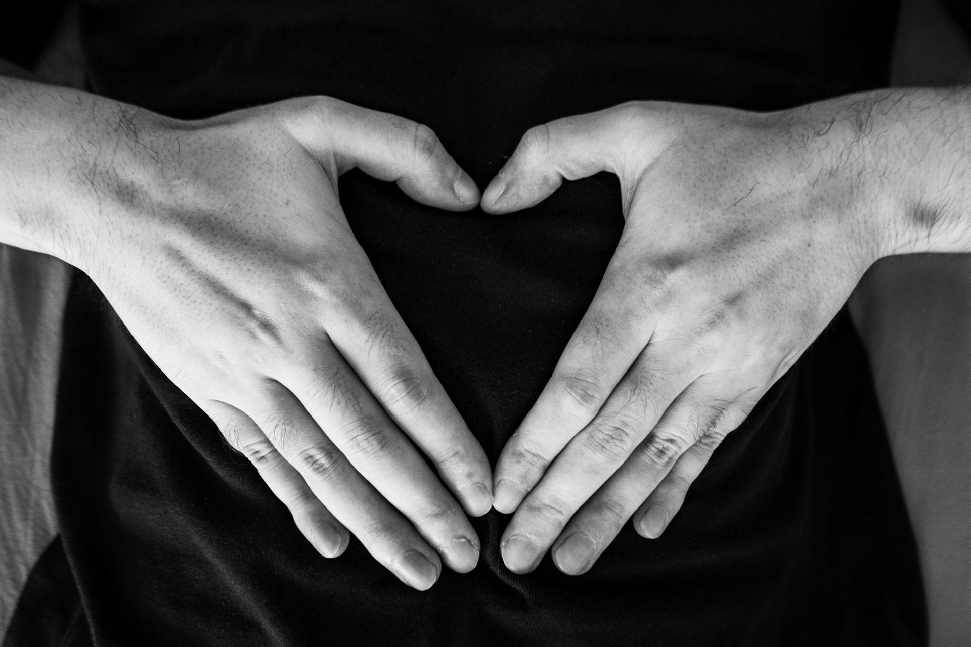 A black-and-white close-up photograph of hands against a dark background, resting on a person’s body. The thumbs and fingers form a heart shape in the middle.