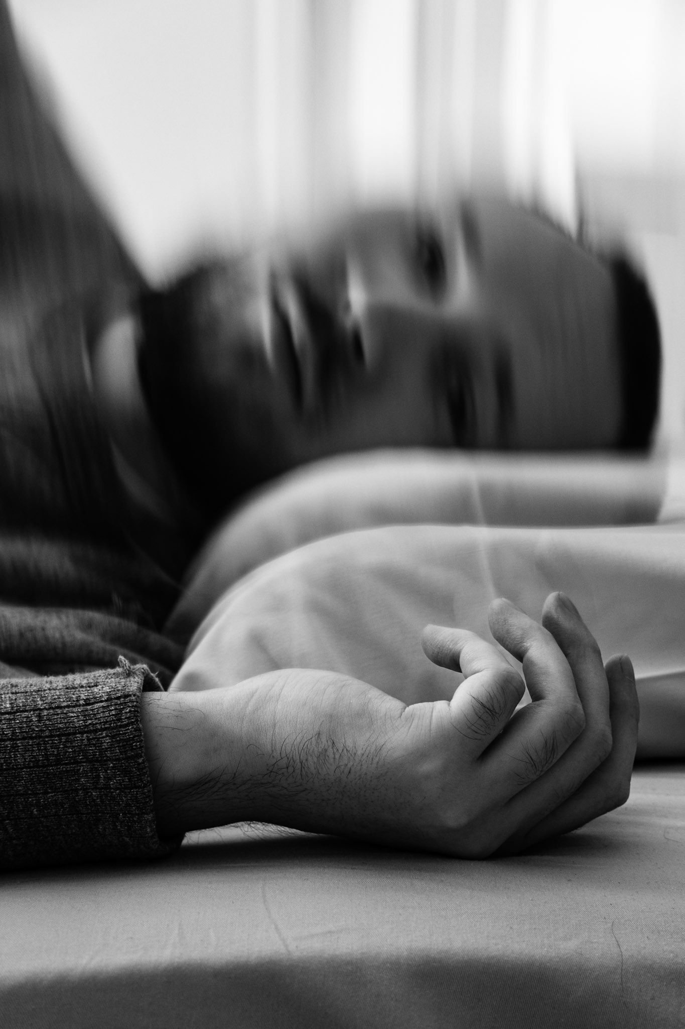A black and white photograph of a man, Niko, resting his head on a pillow. His hand is in the foreground laying against the bed. His face appears blurry and distorted in the background.