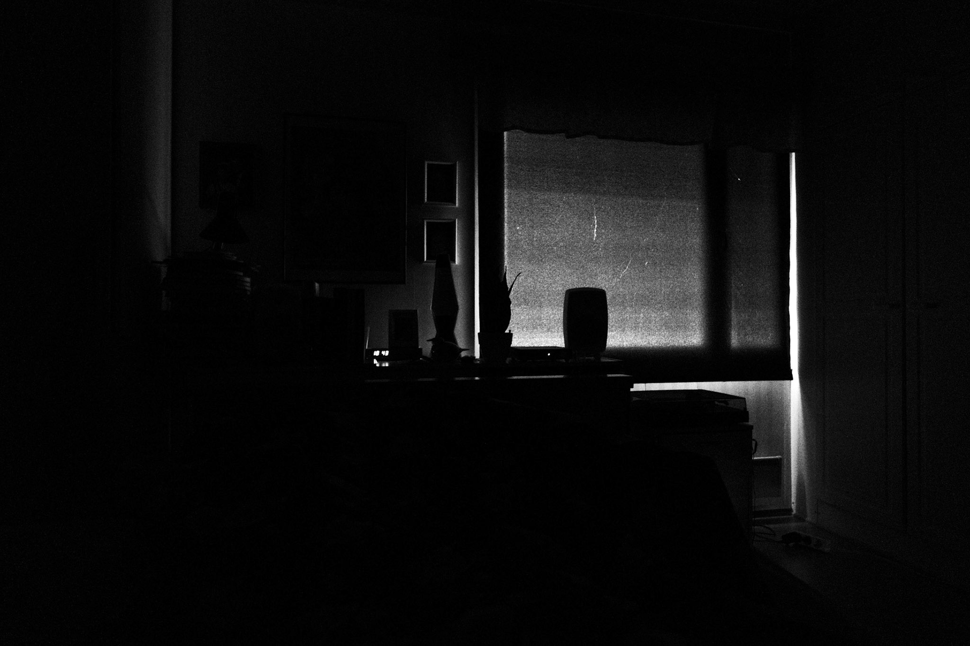A high contrast black and white photograph of a completely dark bedroom. A long and narrow ray of light peaks through the corner of the curtain covered window. The silhouette of the window is seen through the curtain.