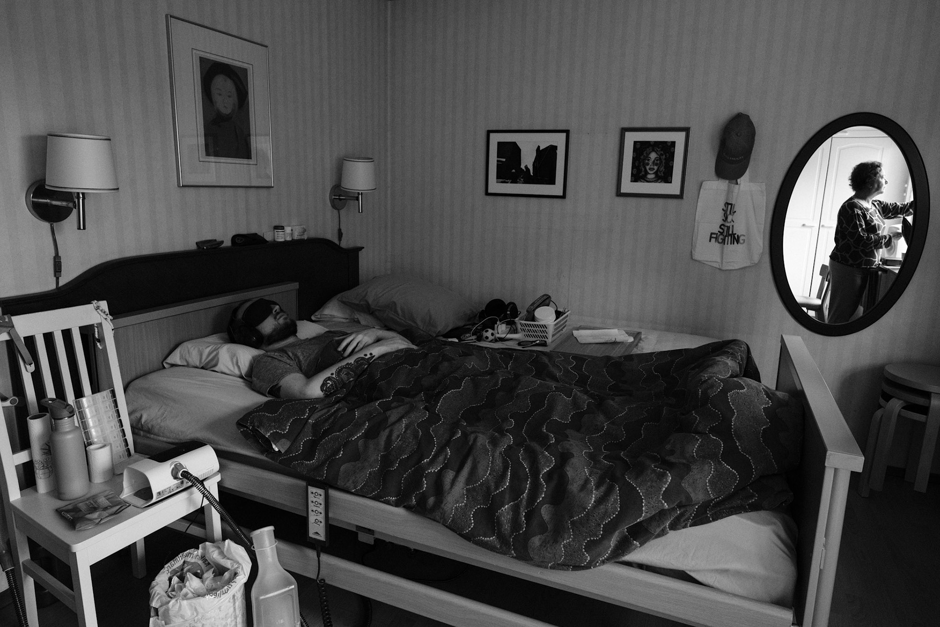 A black and white photo of a man, Niko, lying on a hospital bed, hands crossed on his chest, under a duvet, and wearing an eyemask and earmuffs. The room has framed pictures and personal belongings. A mirror reflects a woman, Päivi, closing the curtains.