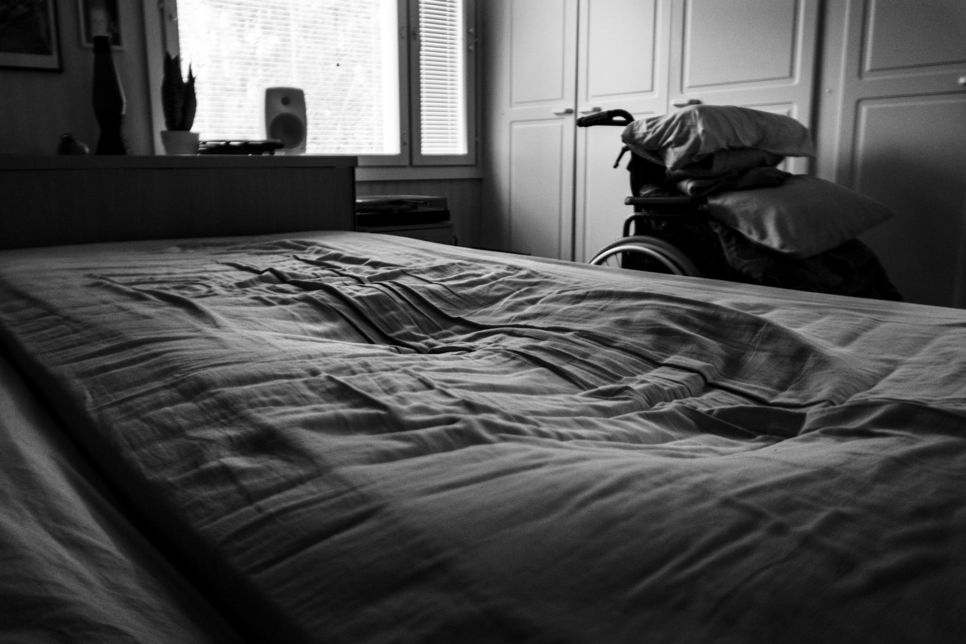 A black-and-white photo of an empty bed, which shows the dip of a human silhouette. A wheelchair, personal belongings, and a window are seen in the background.