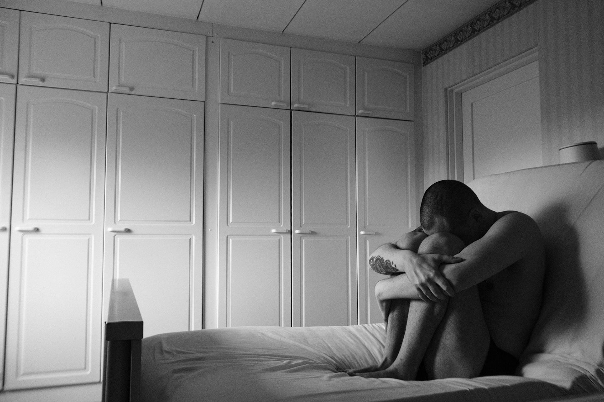 A black and white photograph of a shirtless man sitting on a bed, curled up with his arms wrapped around his knees and his head down. The room has white cabinets in the background, and the setting conveys a sense of isolation. 