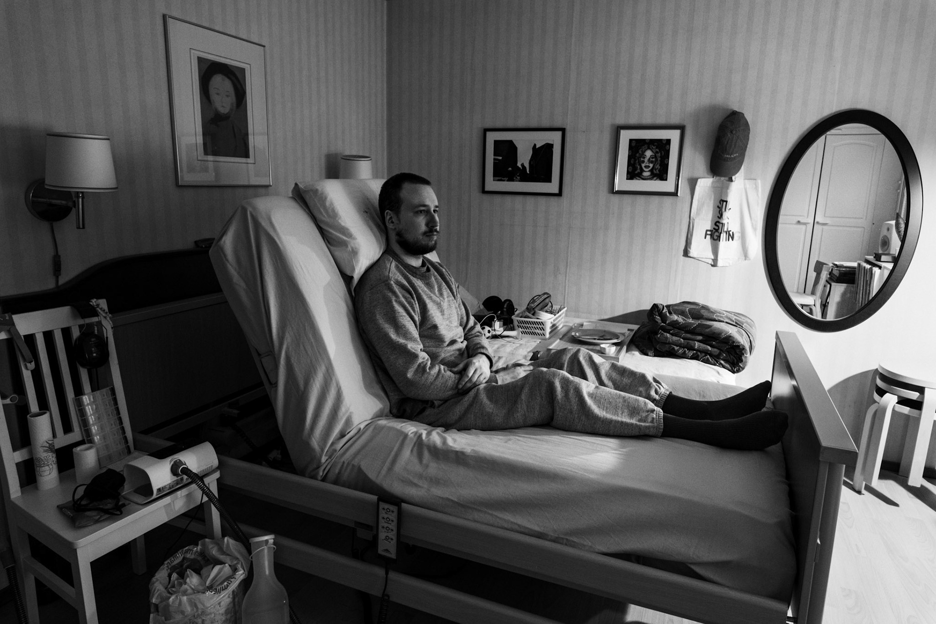 A black-and-white photo of Niko sitting upright in a medical bed, his arms crossed, and he looks out of the window. The room has dim light. A CPAP machine and medical equipment are beside the bed.