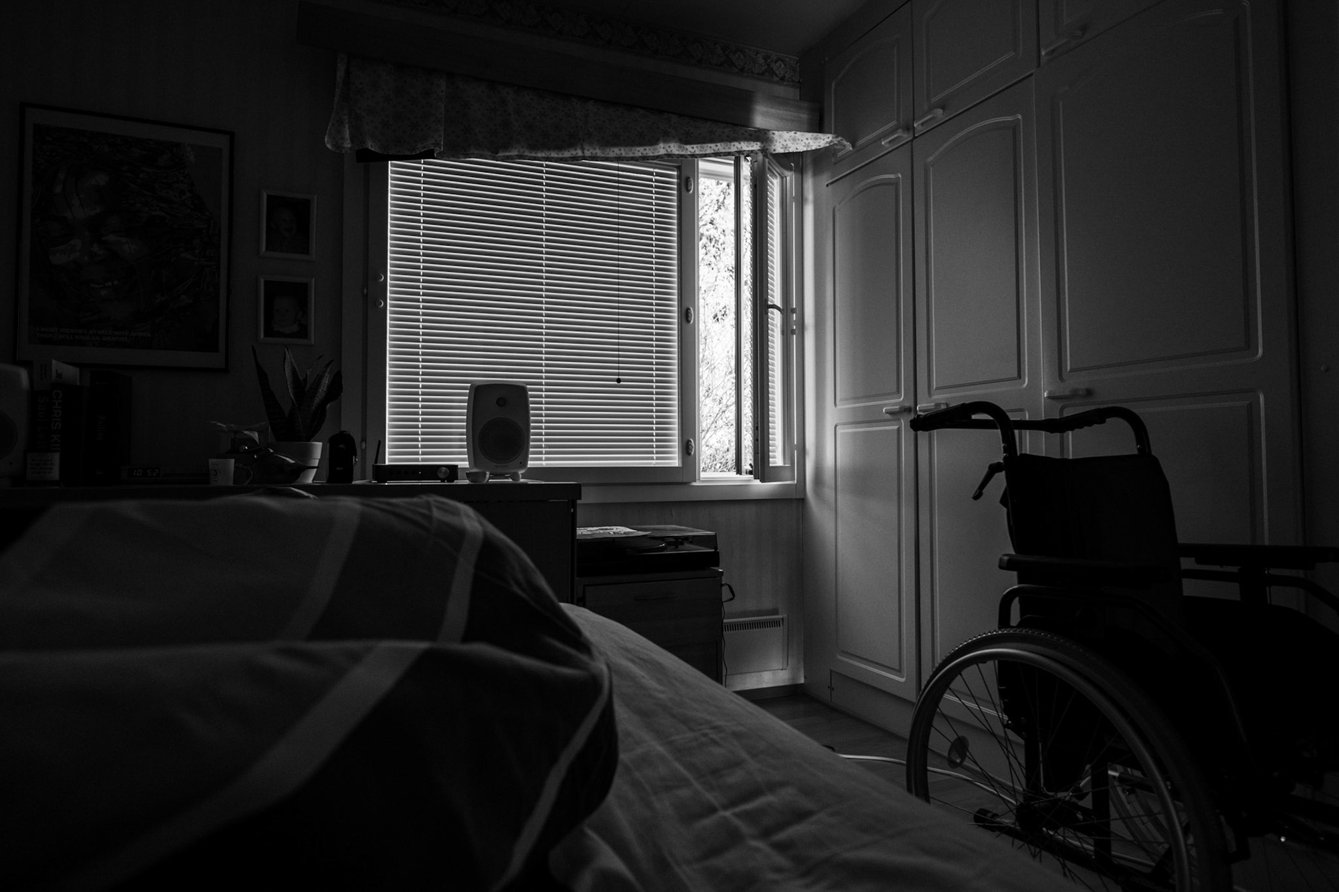 A black-and-white photo taken from Niko’s point of view while he lies in bed. The room is dark; the blinds are closed but the side window is open. A wheelchair is visible next to the bed.