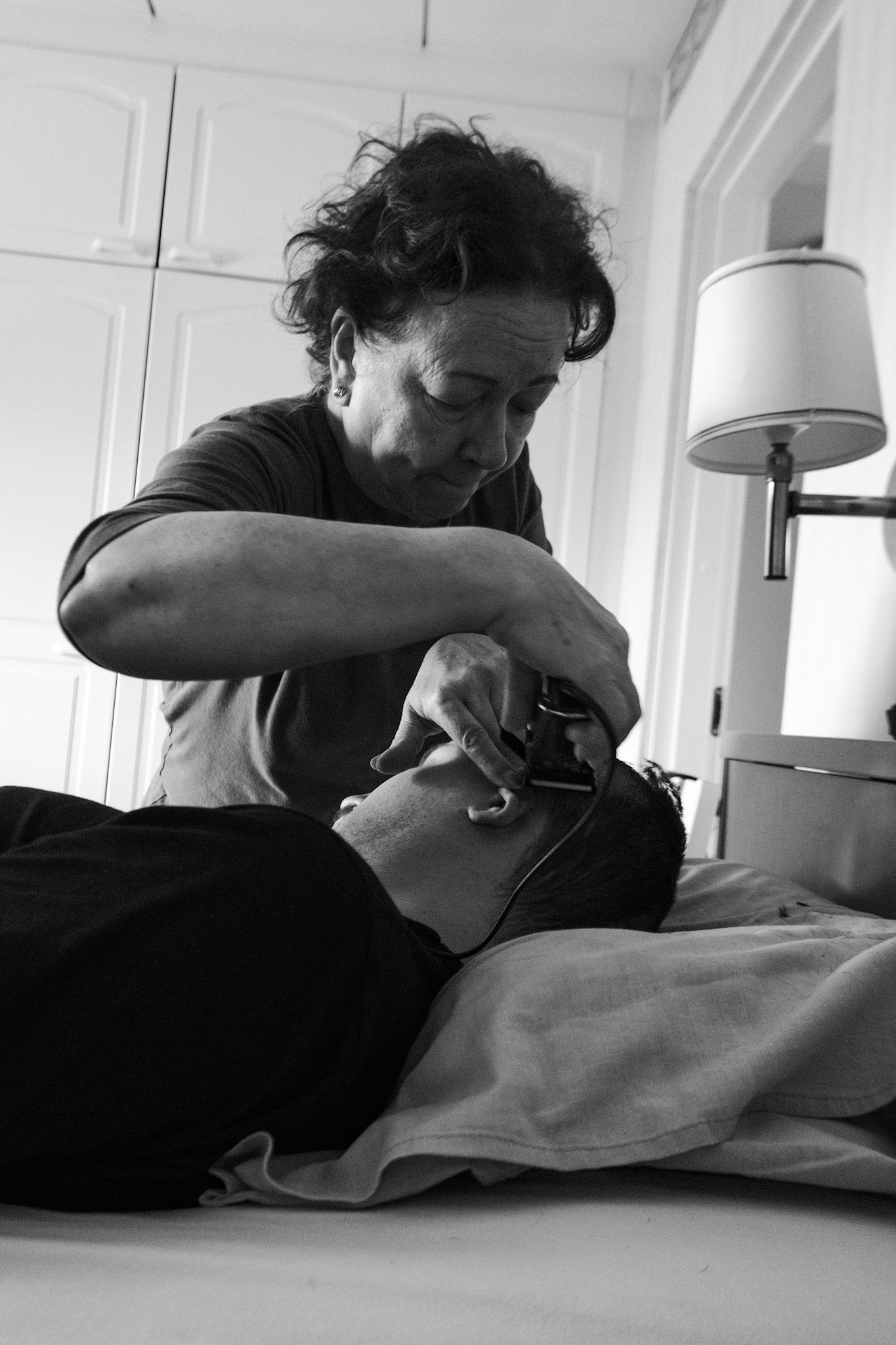 A black and white photo of a woman, Päivi, giving a haircut to Niko, who is lying in a bed. She uses clippers with a focused expression. The bedroom has cabinets in the background and a bedside lamp.