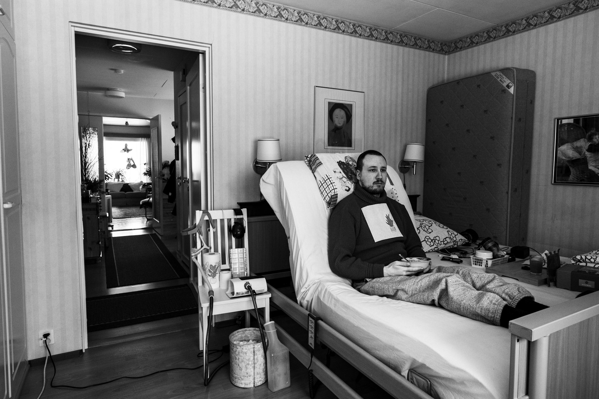 A black-and-white photo of Niko sitting upright in a medical bed, eating a bowl of yoghurt, and looking out of the window. A CPAP machine and medical equipment are beside the bed.