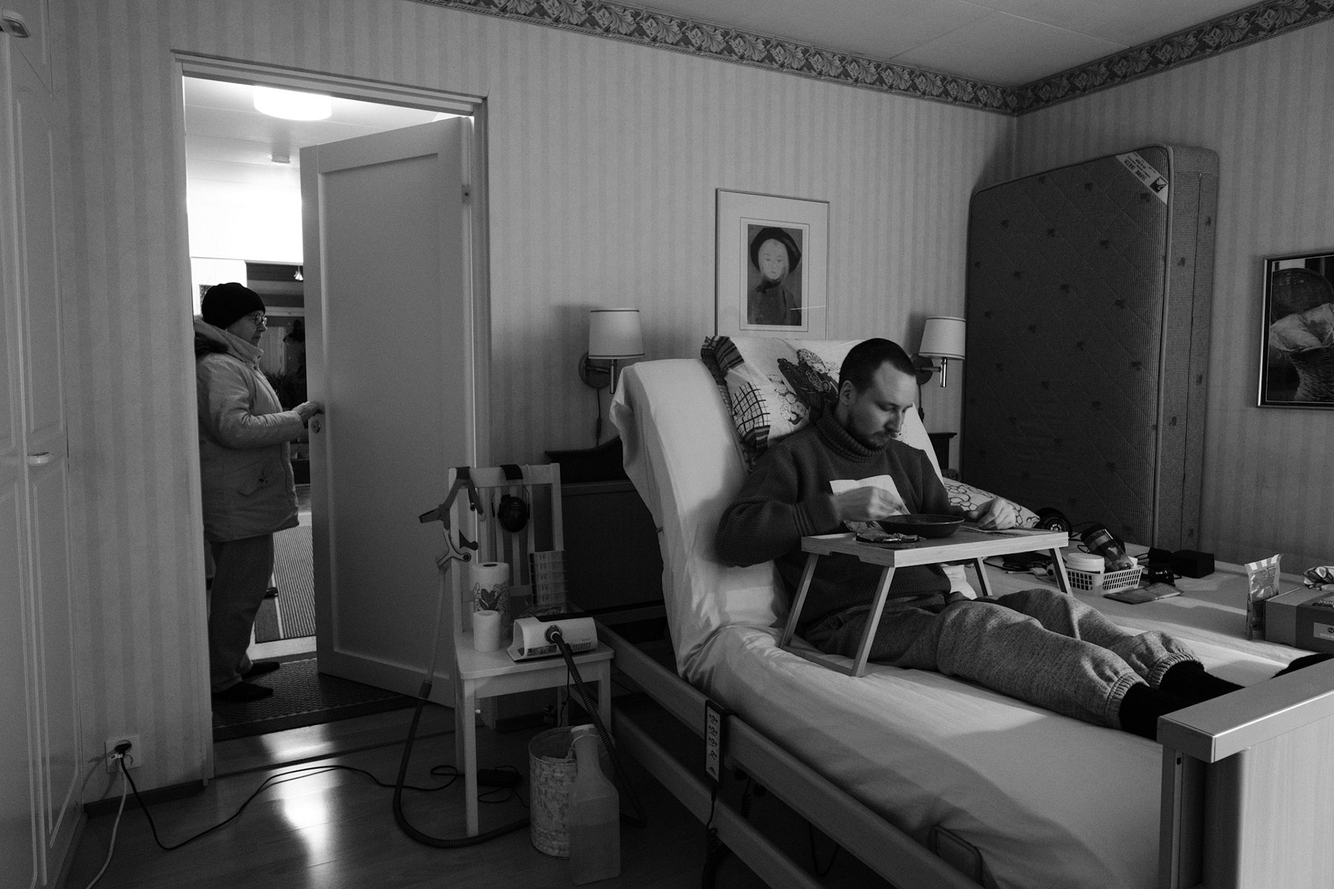 A black and white photo of Niko sitting upright in bed, eating from a tray table while wearing a sweater. Päivi, in a winter coat, stands in the open doorway looking in. A CPAP machine and medical equipment are beside the bed.