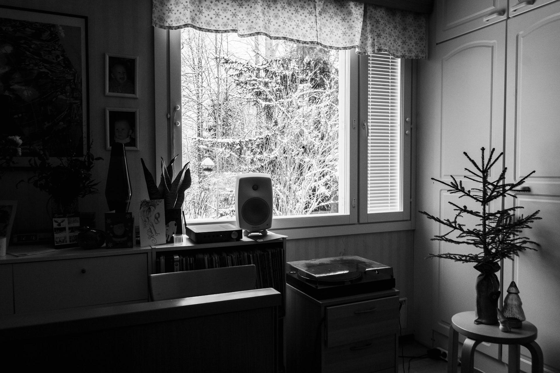 A black-and-white photo of a window view revealing a snowy landscape. Inside the room, there is a small Christmas tree on top of a stool, a turntable and a speaker, and various personal belongings on top of a sideboard.