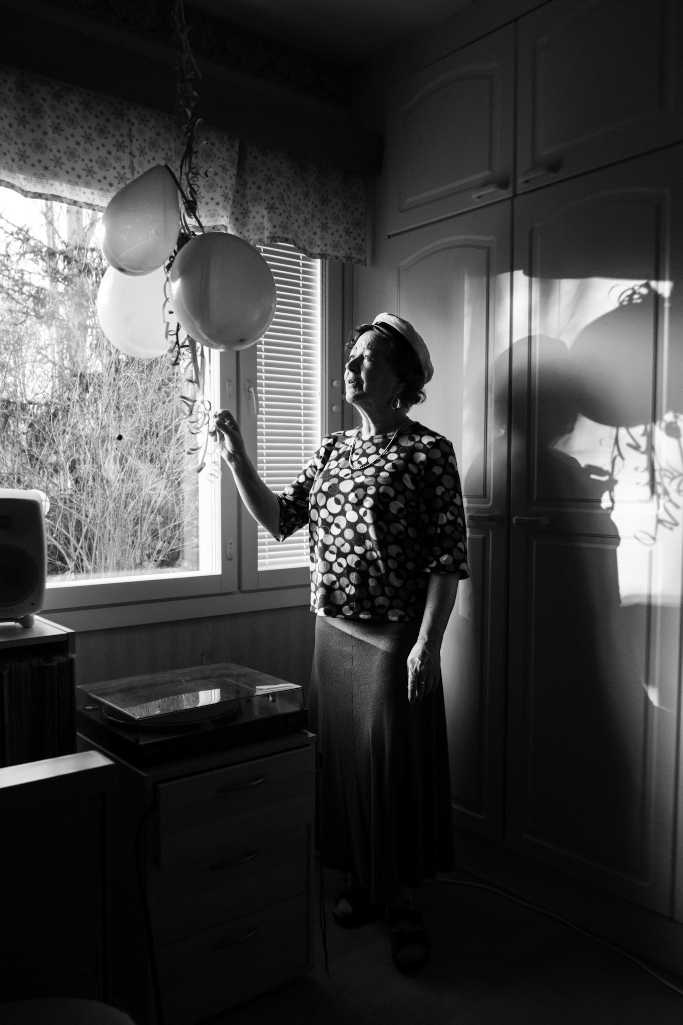 A contrasty black-and-white photograph of a woman in her sixties looking at and reaching her hand towards three balloons that hang from the ceiling in front of a window. She has short curly hair, wears a graduation hat, a blouse and a skirt. A shadow of her shape and the balloons are visible behind her.