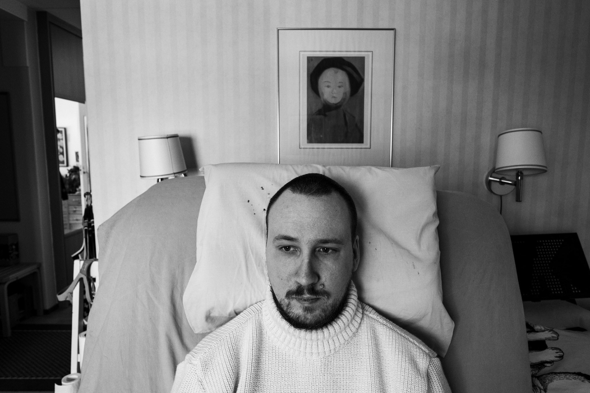 A black-and-white photograph of Niko, a man with a dark beard and a buzz cut, wearing a turtleneck jumper while sitting on his hospital bed. He looks just outside the camera. His bedroom, a small section of a hallway, and a painting resembling his shape are visible in the background.