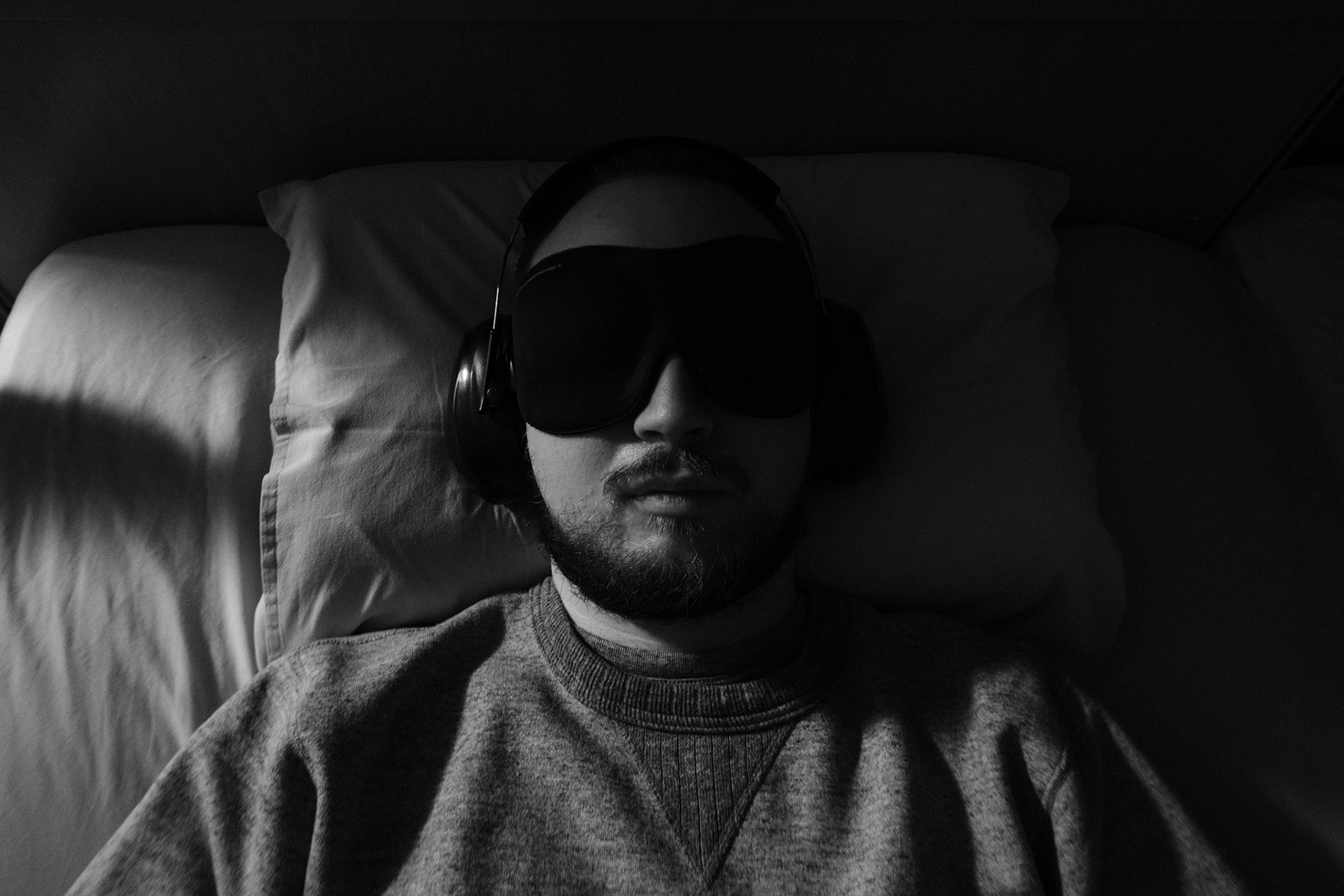A black and white photo of a man with a dark beard and short hair, Niko, lying in a dark bedroom, in bed with his eyes closed. He is wearing an eyemask and earmuffs. The light comes from the side and lits half of his face.