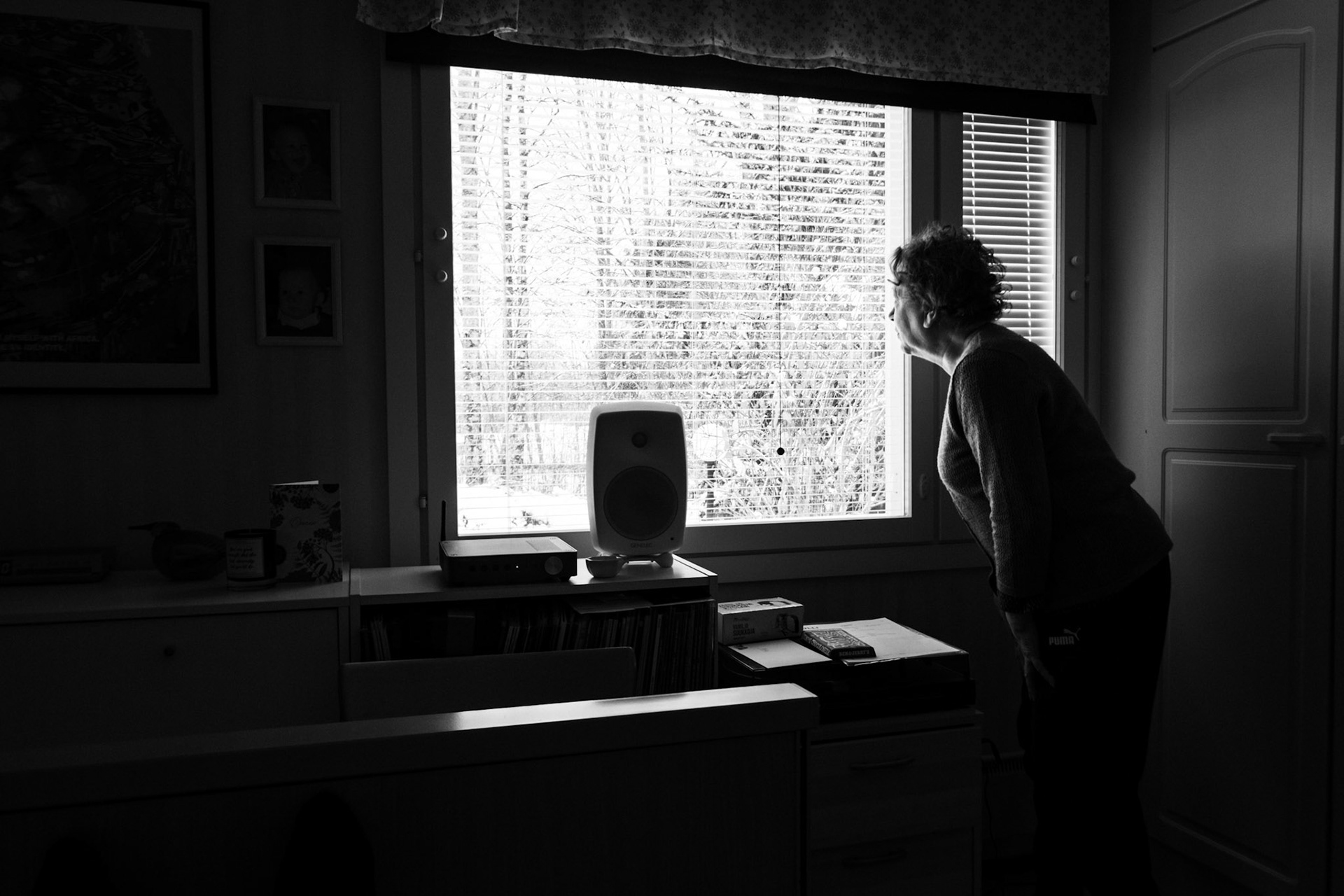 A black-and-white photograph of a woman, Päivi, standing inside a dimly lit room, leaning forward to look out of a window. The window reveals a snowy landscape outside, while the room is filled with furniture, books, and various objects.