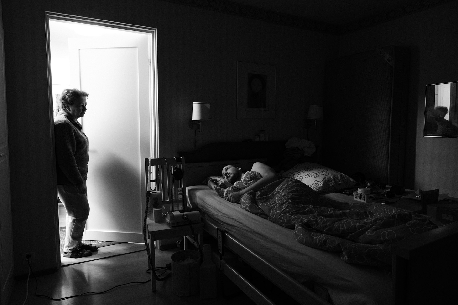 A black and white photo of a dimly lit bedroom. Niko, a man with a shaved head and beard, lies in bed under a blanket, looking at Päivi, his mother, who stands in the doorway, lit by another room. A chair beside the bed holds medical equipment, personal items.