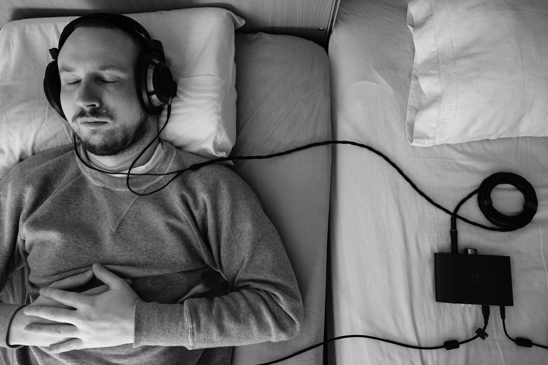 A black and white photo of a man, Niko, with short hair and a beard, wearing a grey sweatshirt, resting in bed and listening to music eyes closed from wired headphones. Headphones are connected to a DAC/Amp. His hands resting on his chest.