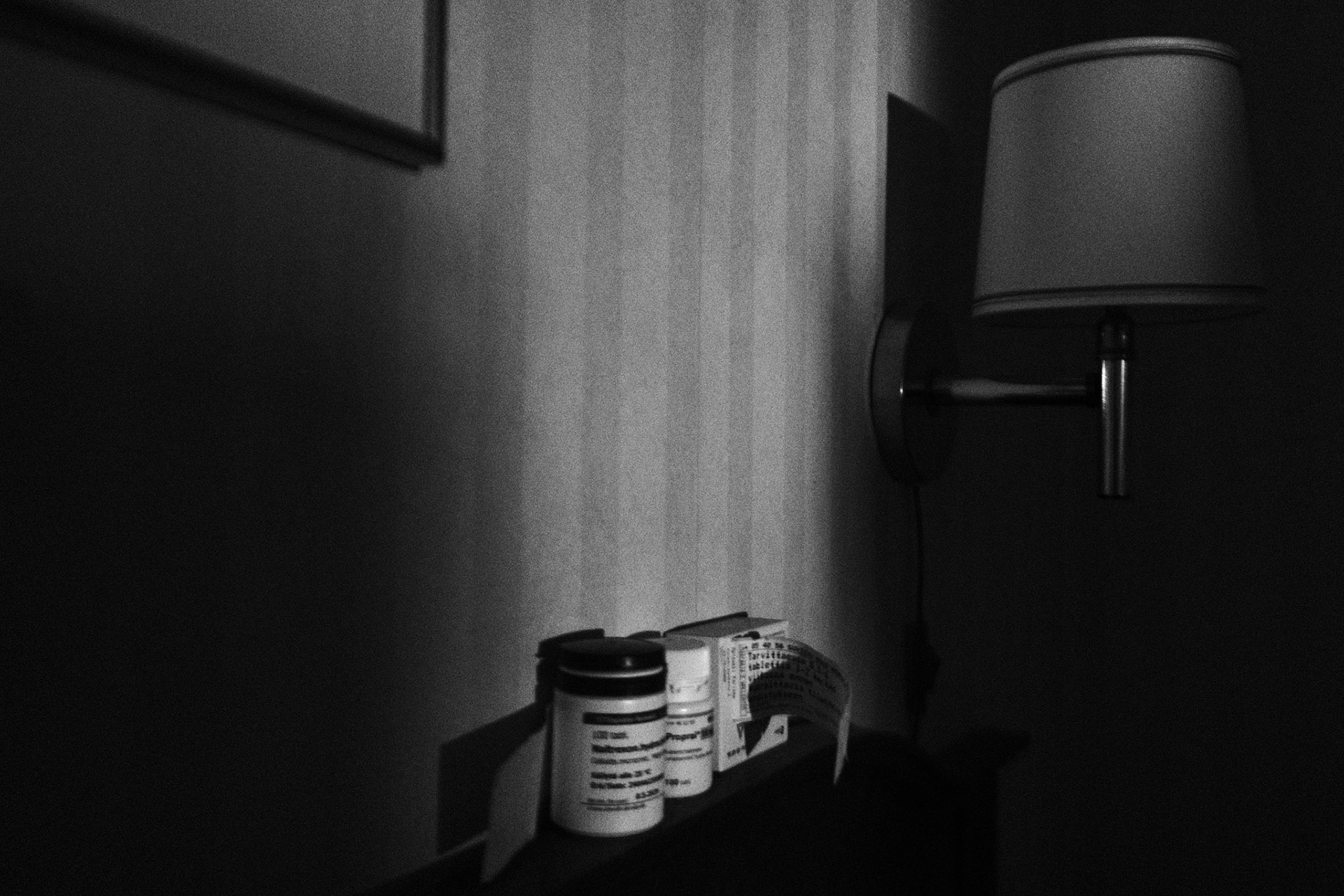 A black and white photo of three medication bottles on top of a bedframe, the light illuminates the bottles in an otherwise dark bedroom. A wall lamp and a frame is also part of the picture.