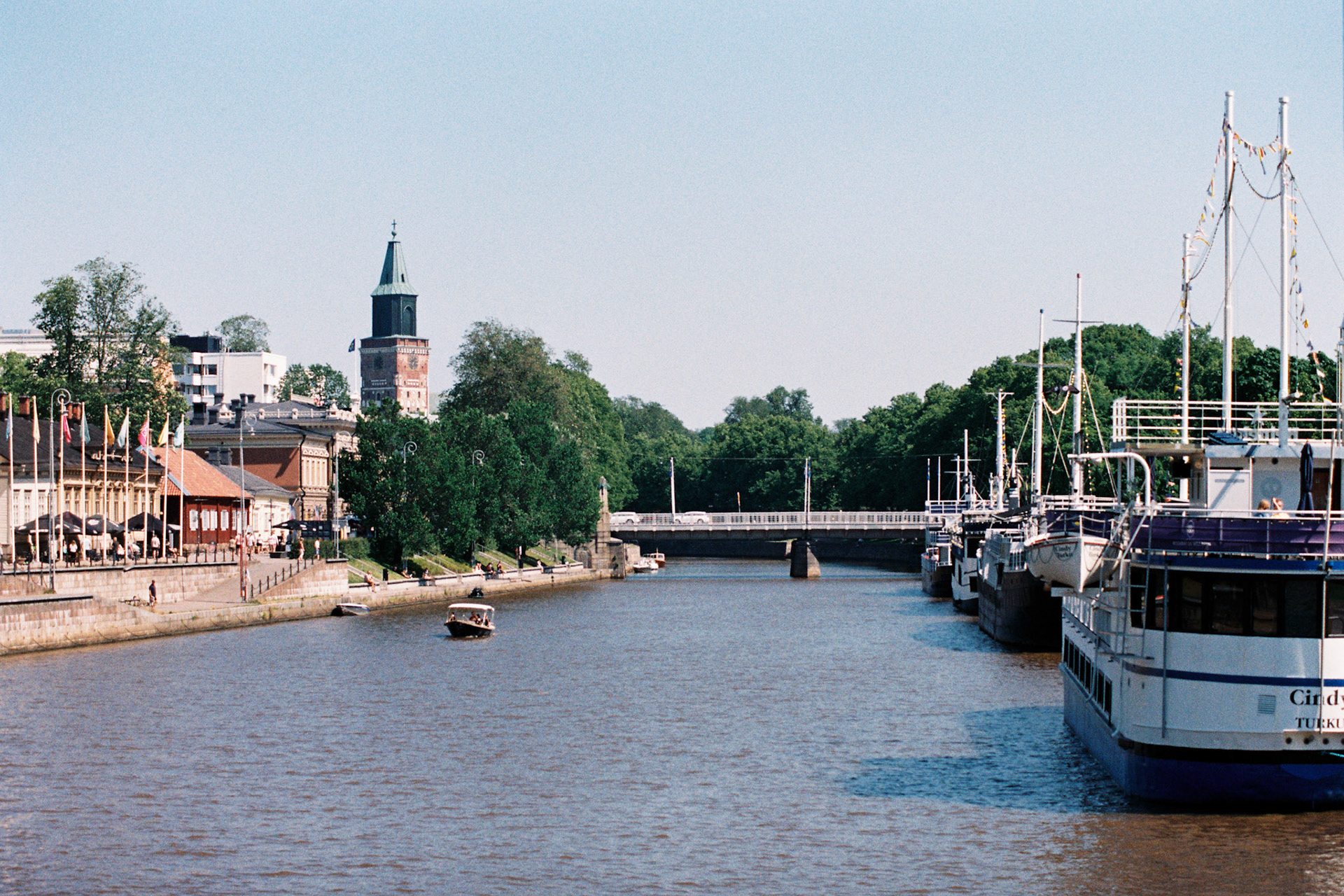A colour photograph of the river Aurajoki in Turku. Tuomiokirkko, the church, is visible in the distance. Big restaurant ships are docked next to the riverbank. A small motorboat is moving across the river