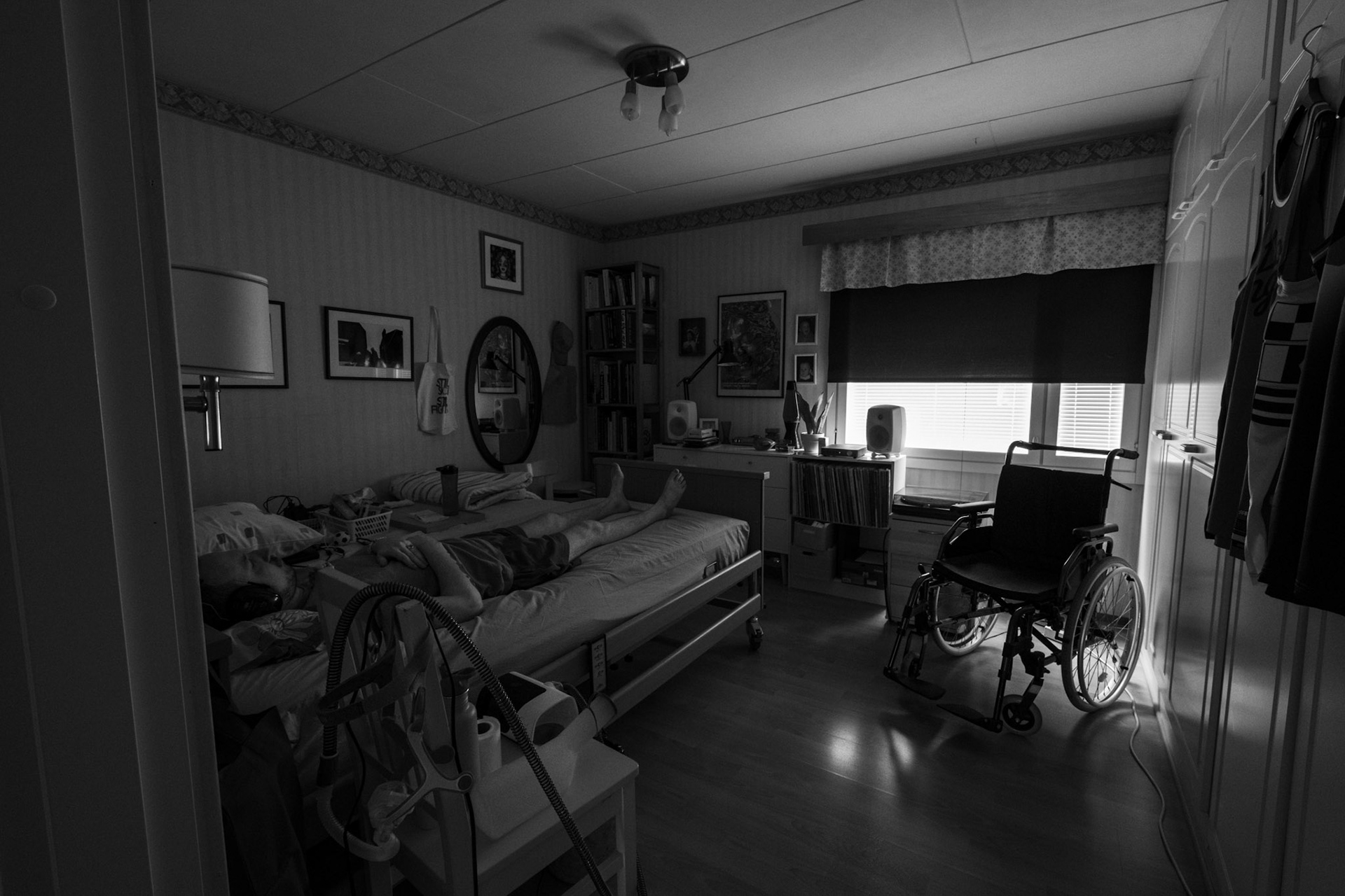 A black-and-white photograph of a dark bedroom where Niko is lying on his bed eyes closed while he wears earmuffs. The blinds of the window are shut, and the blackout curtain is lowered. A wheelchair is in front of the window. The room has artworks and an oval-shaped mirror hanging on the walls. A chair with personal objects is next to the bed.