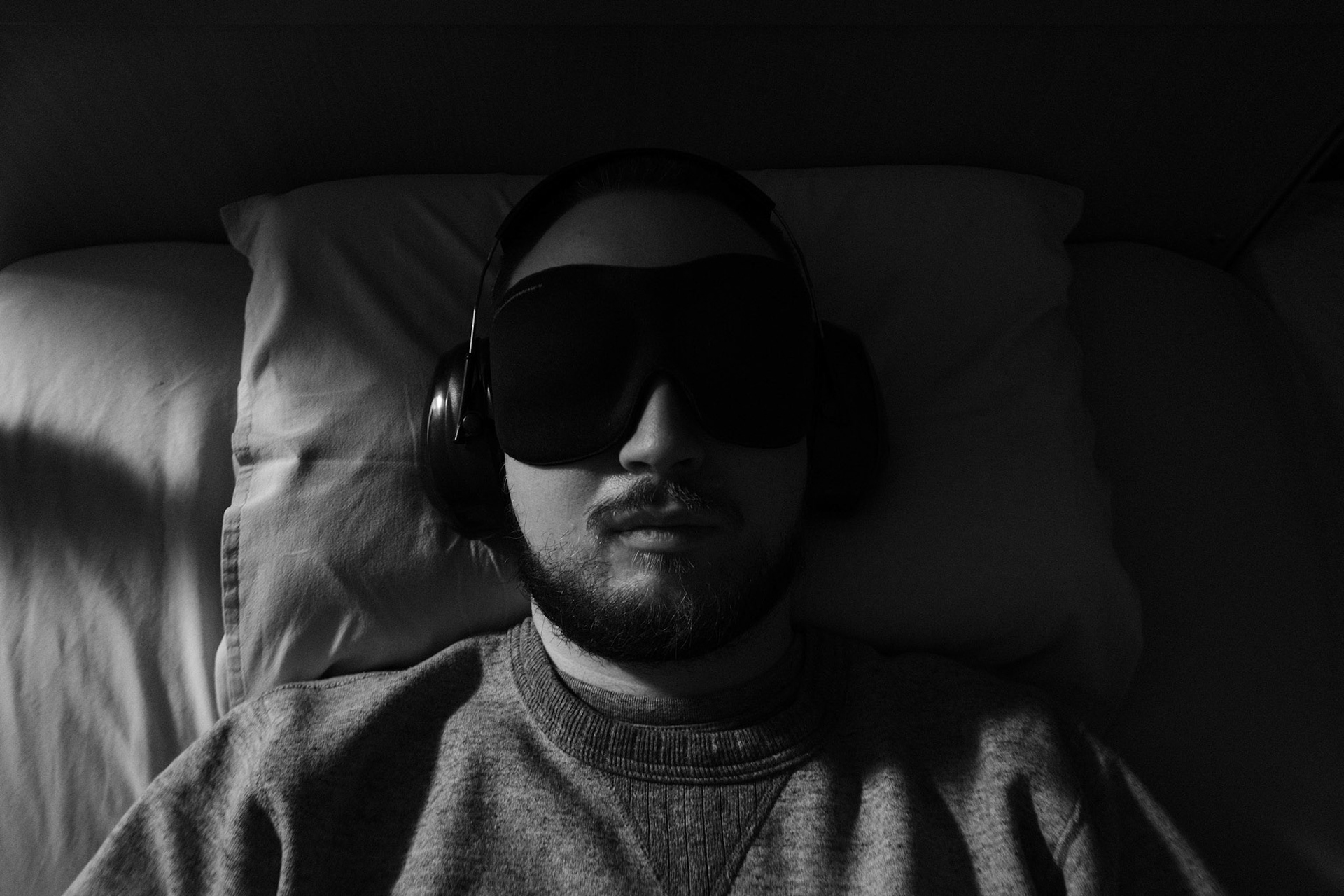 A black and white photo of a man with a dark beard and short hair, Niko, lying in a dark bedroom, in bed with his eyes closed. He is wearing an eyemask and earmuffs. The light comes from the side and lits half of his face.