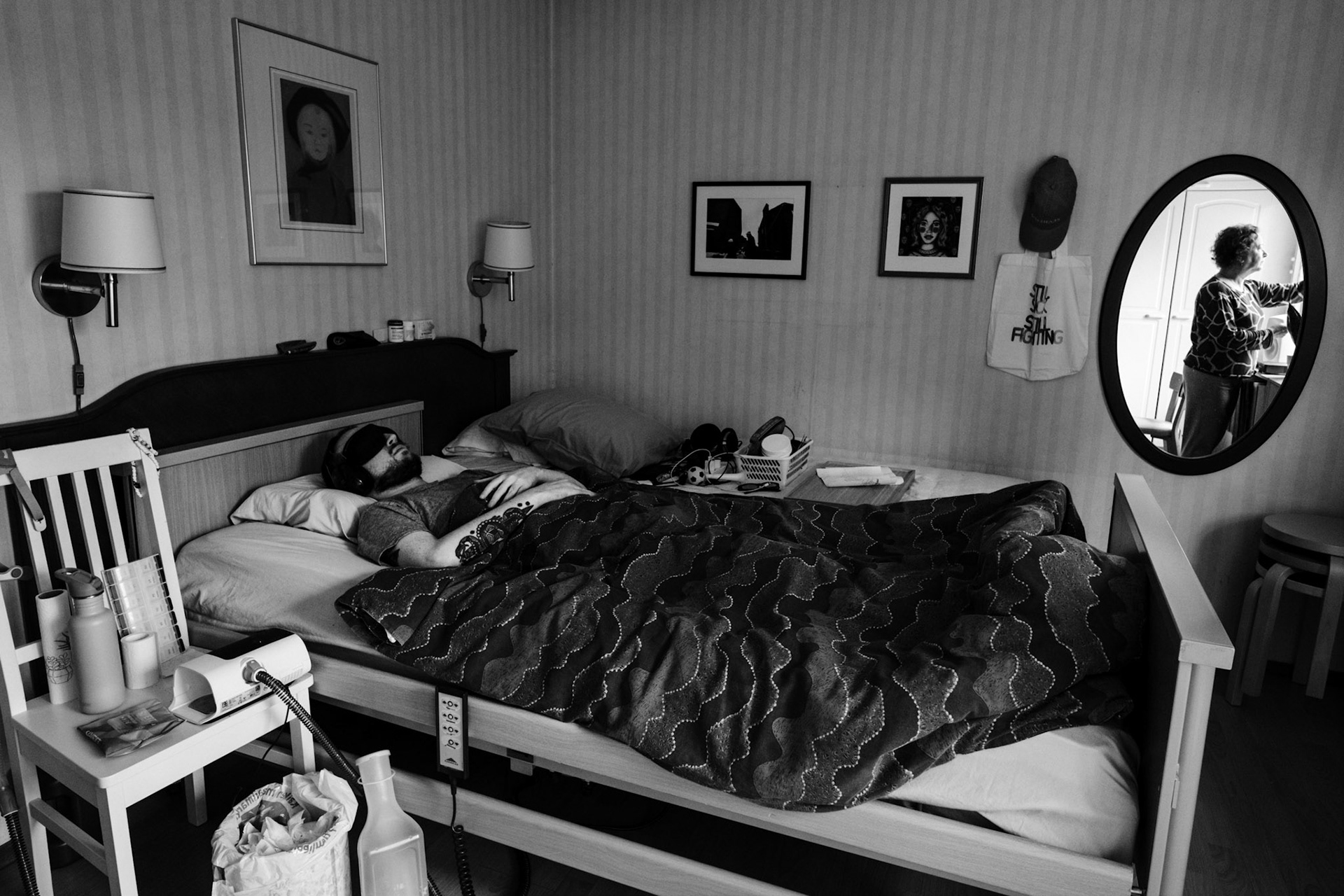 A black-and-white photo of a man, Niko, lying on a hospital bed, hands crossed on his chest, under a duvet, and wearing an eyemask and earmuffs. The room has framed pictures and personal belongings. A mirror reflects a woman, Päivi, closing the curtains.