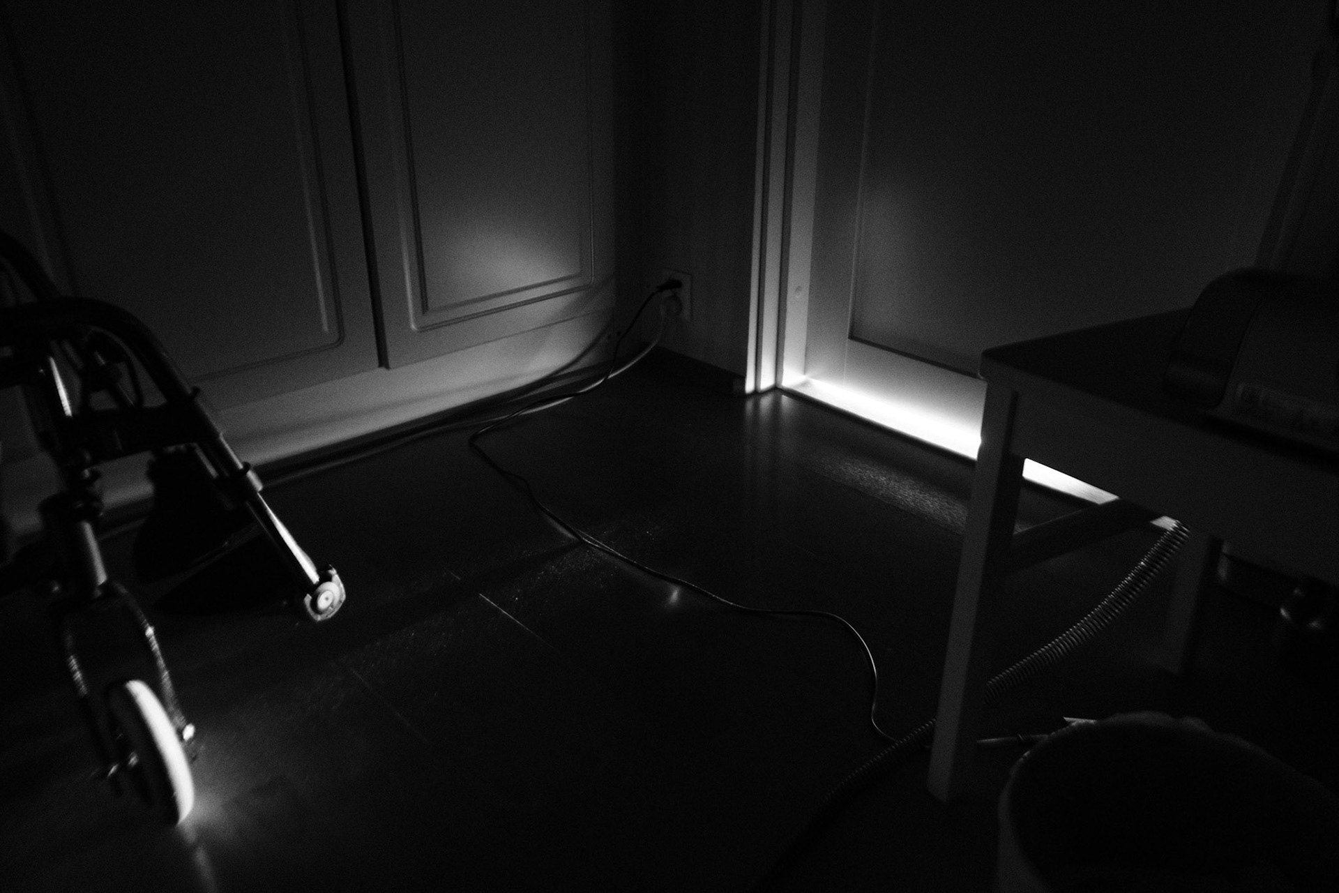 A black and white photograph of a dark bedroom floor. The light peeks through under the  closed door, lits part of a wheelchair and a cable running across the floor.