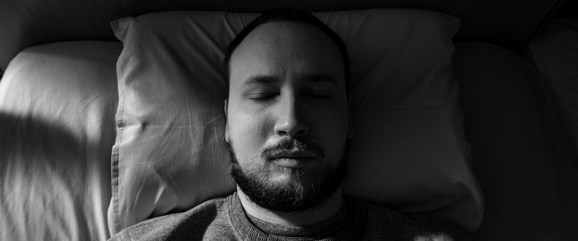  A black-and-white photo of a man with a dark beard and short hair, Niko, lying in a dark bedroom, in bed with his eyes closed. The light comes from the side and lits half of his face.