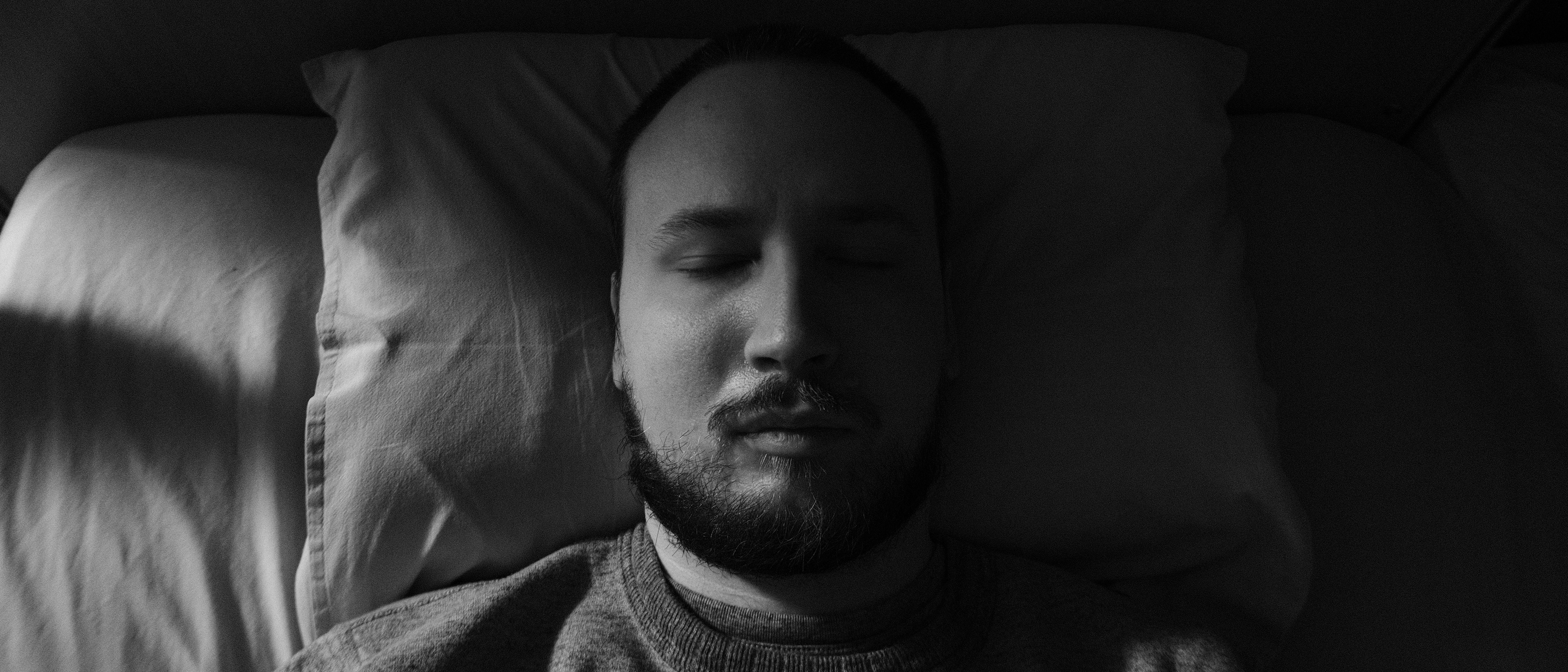A black and white photo of a man with a dark beard and short hair, Niko, lying in a dark bedroom, in bed with his eyes closed. The light comes from the side and lits half of his face.