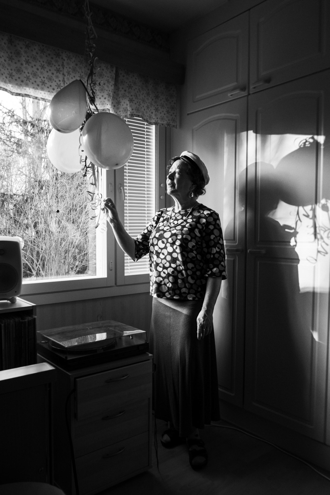 A contrasty black-and-white photograph of a woman in her sixties looking at and reaching her hand towards three balloons that hang from the ceiling in front of a window. She has short curly hair, wears a graduation hat, a blouse and a skirt. A shadow of her shape and the balloons are visible behind her.
