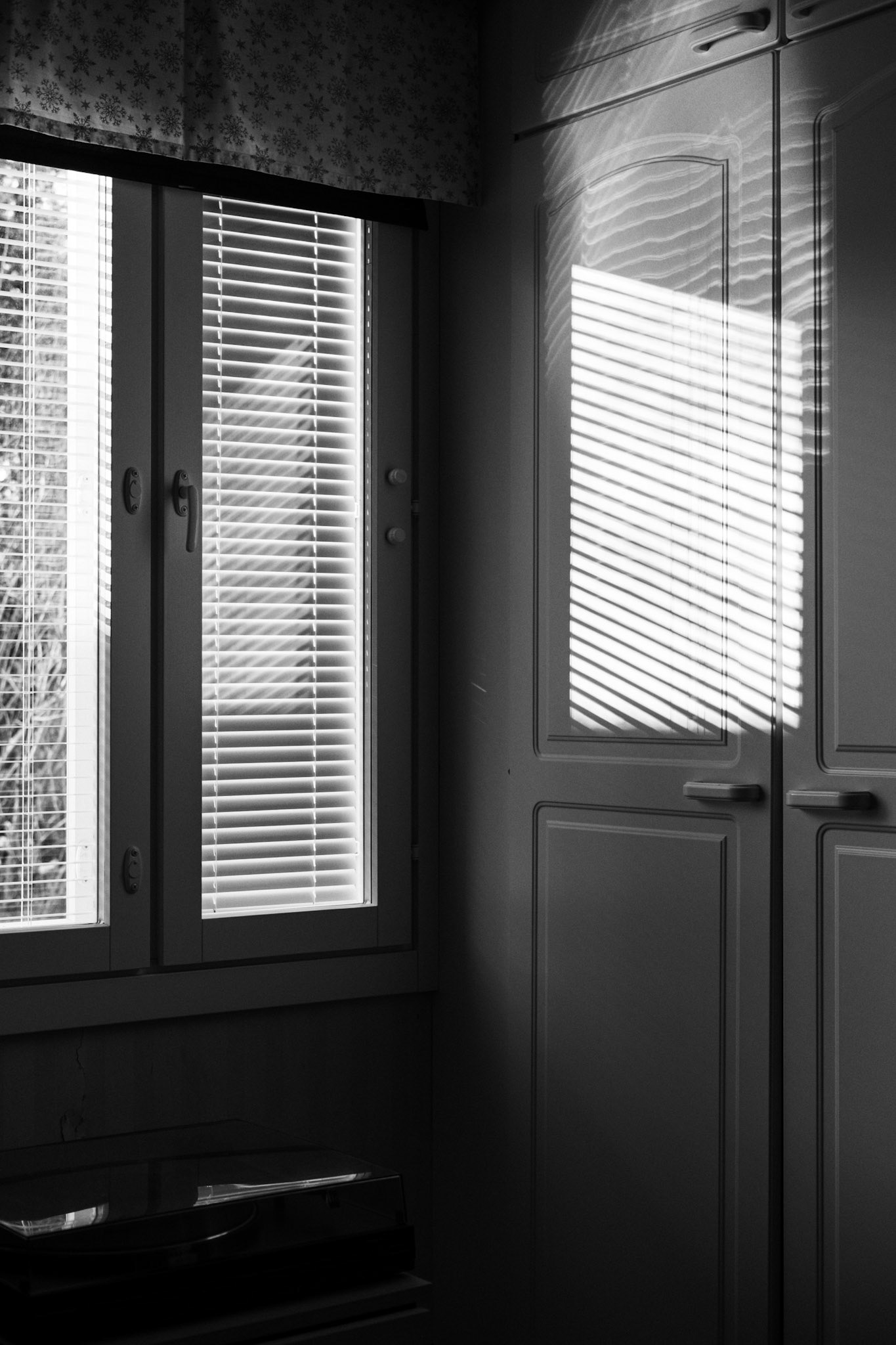A black-and-white photograph of a bedroom corner: an abstract pattern of light and shadow forms on a closet’s door next to a window.