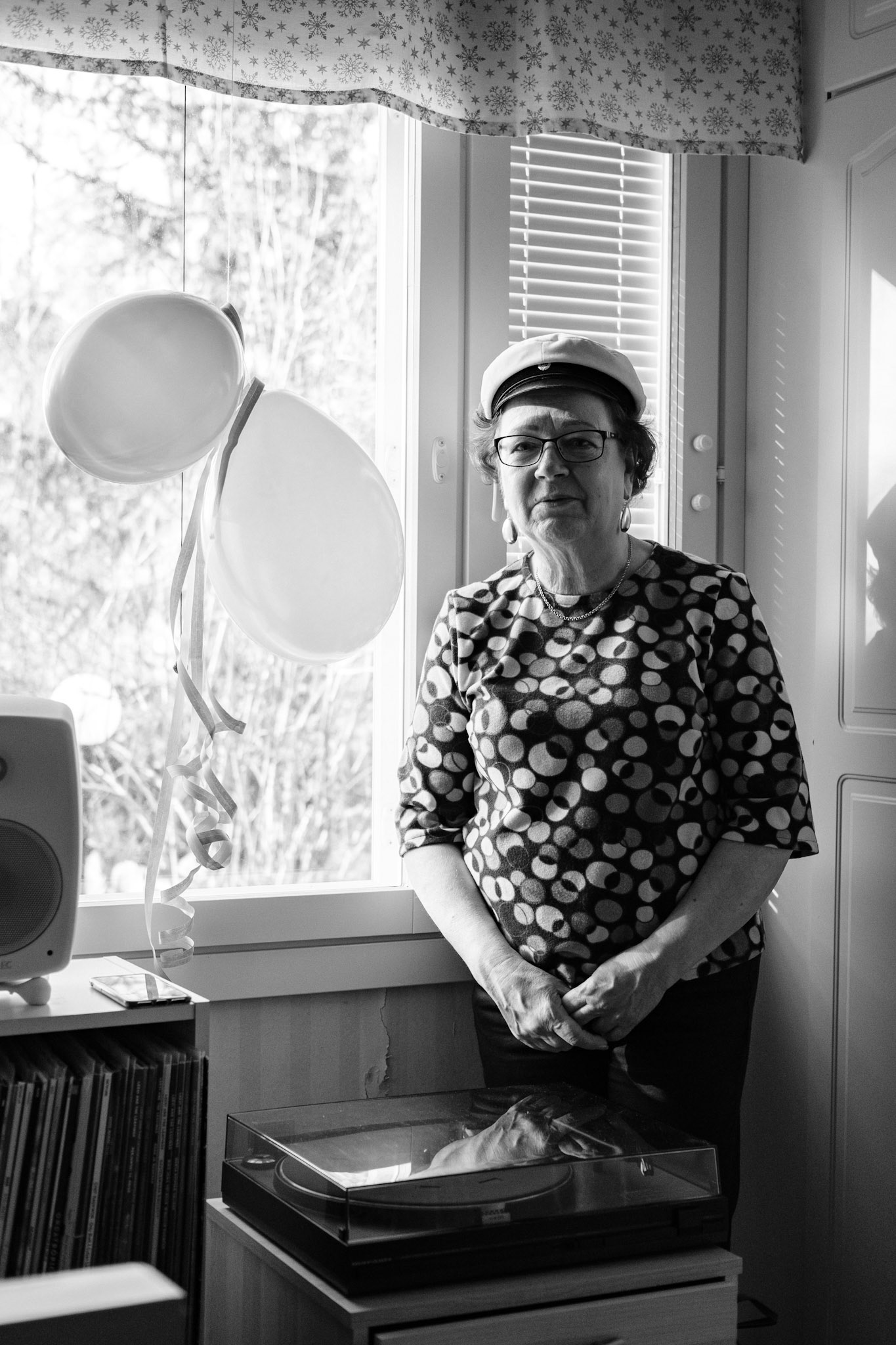 A black and white photo of Niko’s mother wearing her graduation hat and standing next to a window. Two balloons and ribbons hang in front of the window.