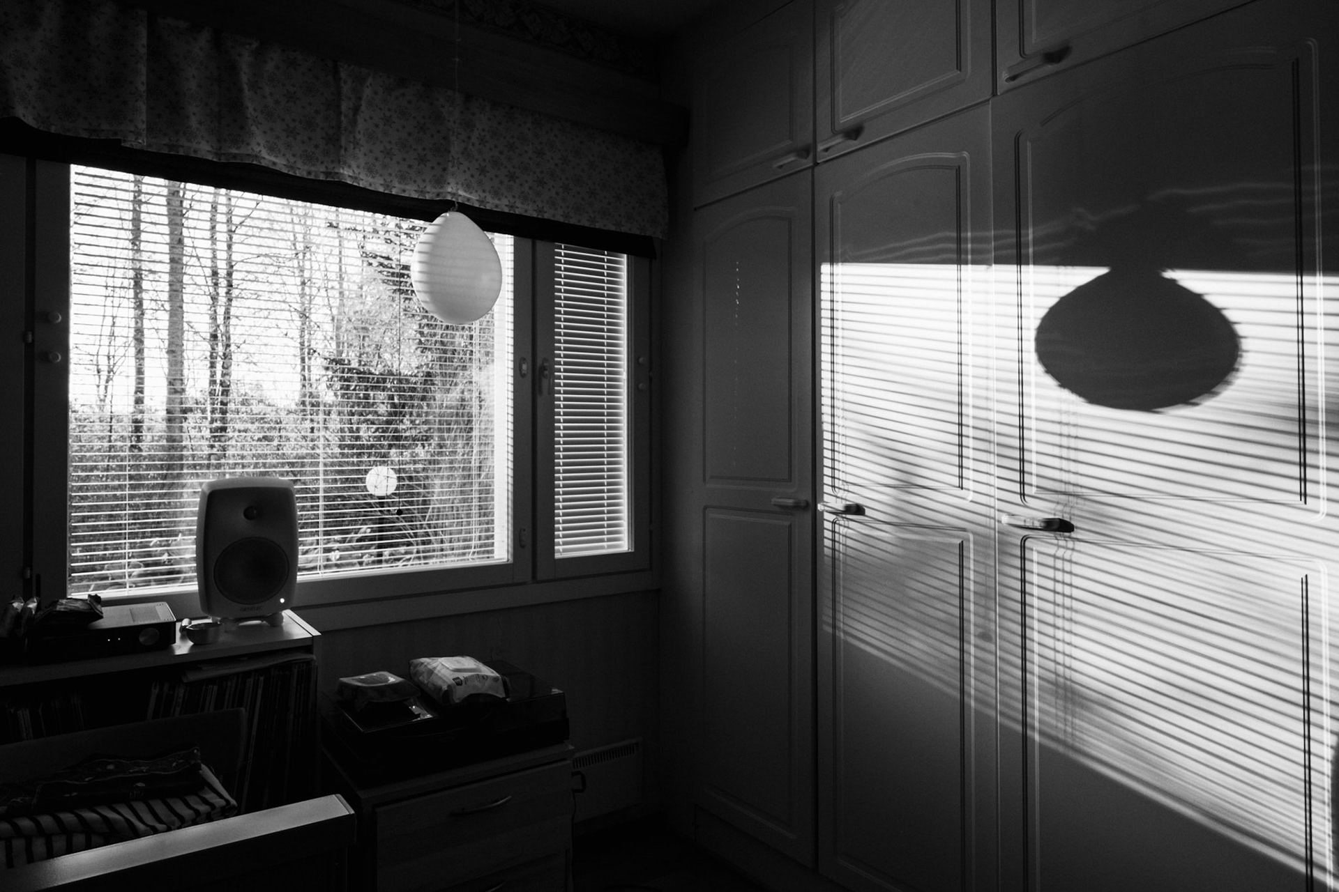 A black-and-white photo of a window view revealing a spring landscape. Inside the room, there is a balloon hanging in front of the window, which casts a long shadow onto a closet. There is also a turntable and a speaker, and various personal belongings on top of a sideboard.