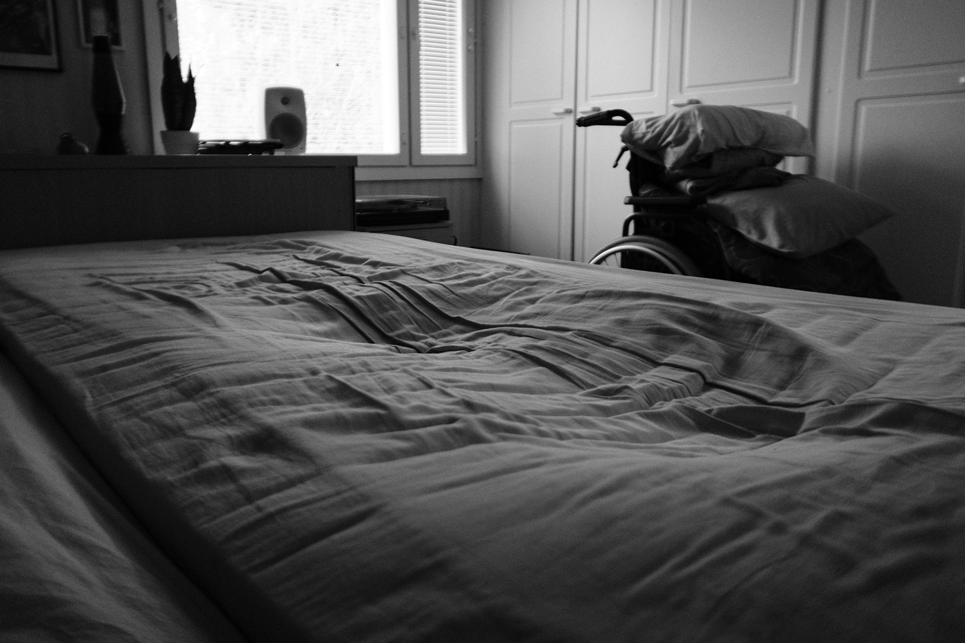 A black and white photo of an empty bed, which shows the dip of a human silhouette. A wheelchair, personal belongings, and a window are seen in the background.