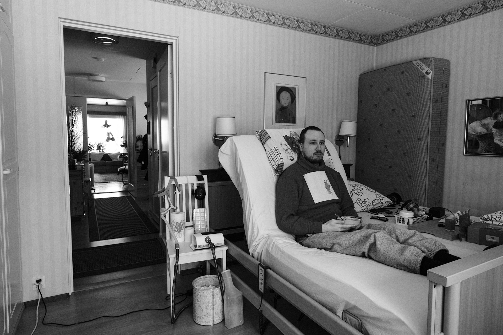 A black and white photo of Niko sitting upright in a medical bed, eating a bowl of yoghurt, and looking out of the window. A CPAP machine and medical equipment are beside the bed.