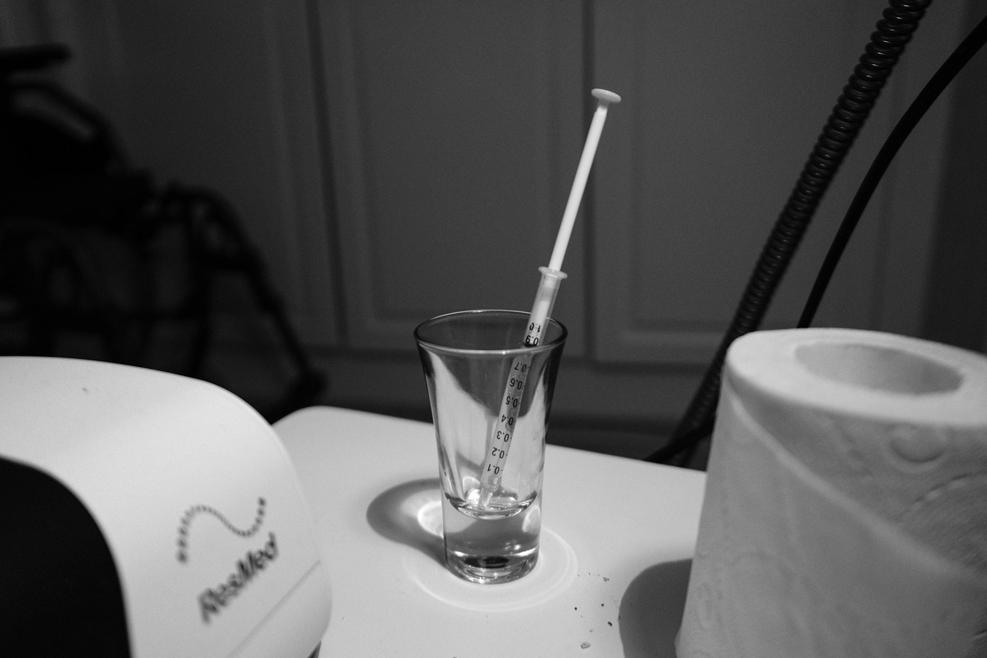 A black and white photo of a medication syringe full of liquid medication, placed in a shot glass. In the foreground are a roll of toilet paper and a CPAP machine.