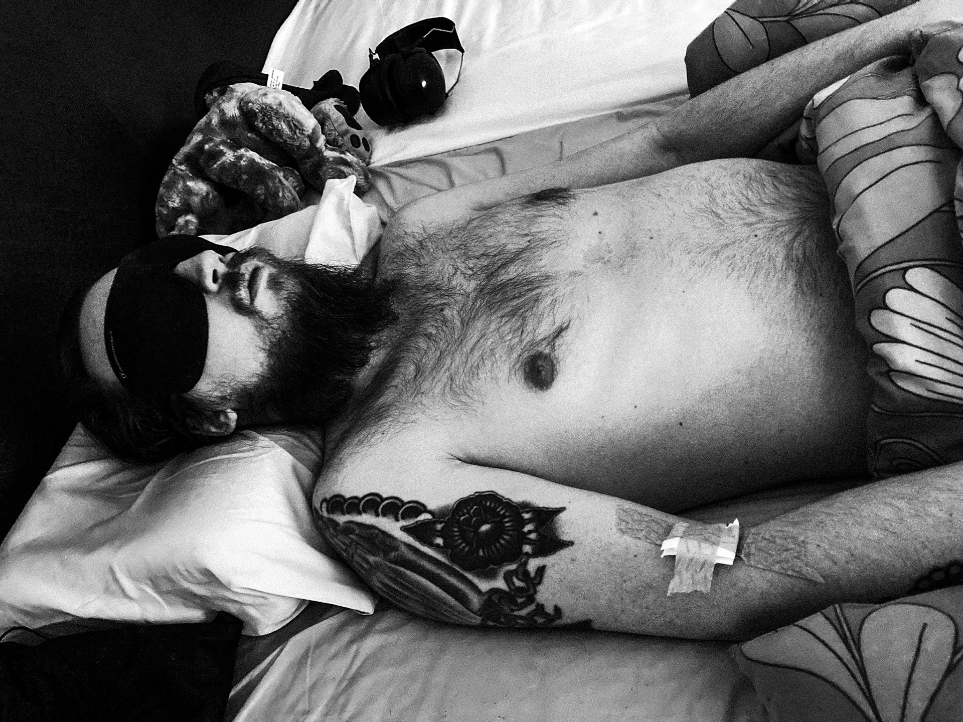 A black-and-white photo of a shirtless Niko, with a messy long beard, lying in bed with an eye mask on. A bandage on his right arm. Ear protectors are lying next to him.