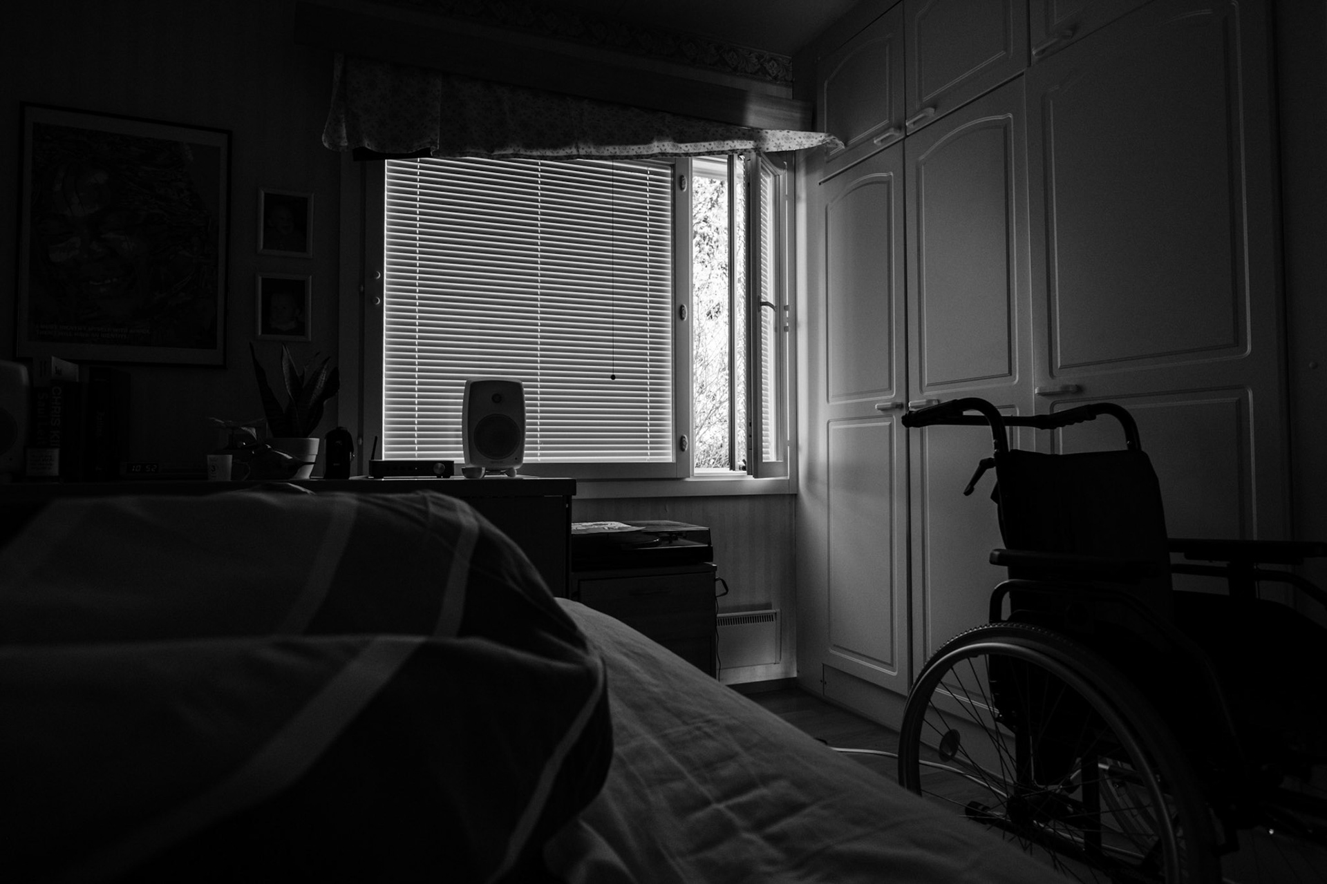 A black-and-white photo taken from Niko’s point of view while he lies in bed. The room is dark; the blinds are closed but the side window is open. A wheelchair is visible next to the bed.
