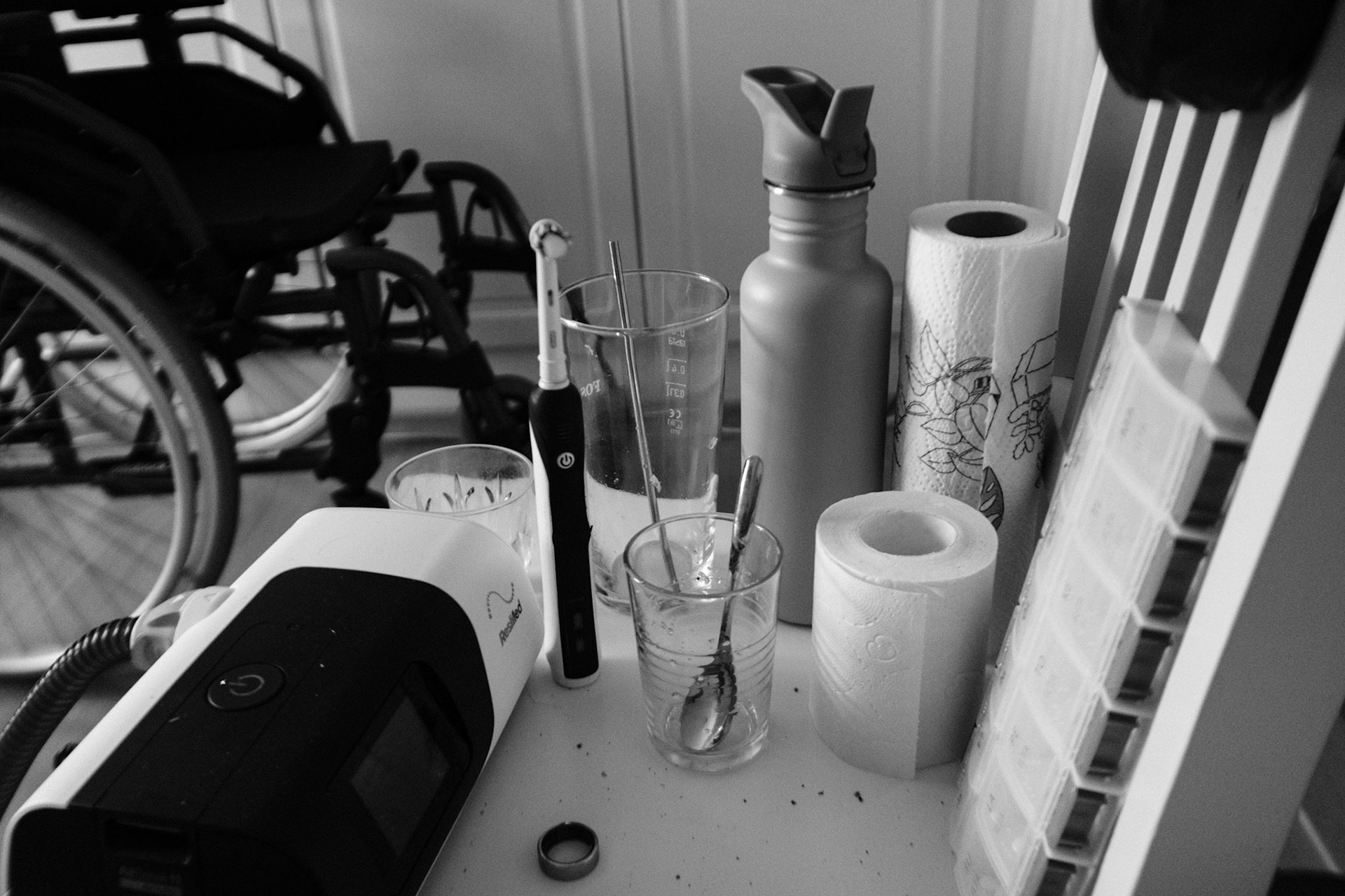 A black and white photo of a cluttered chair with an electric toothbrush, water bottle, paper towel roll, pill organizer, and other personal items. A wheelchair is in the background.