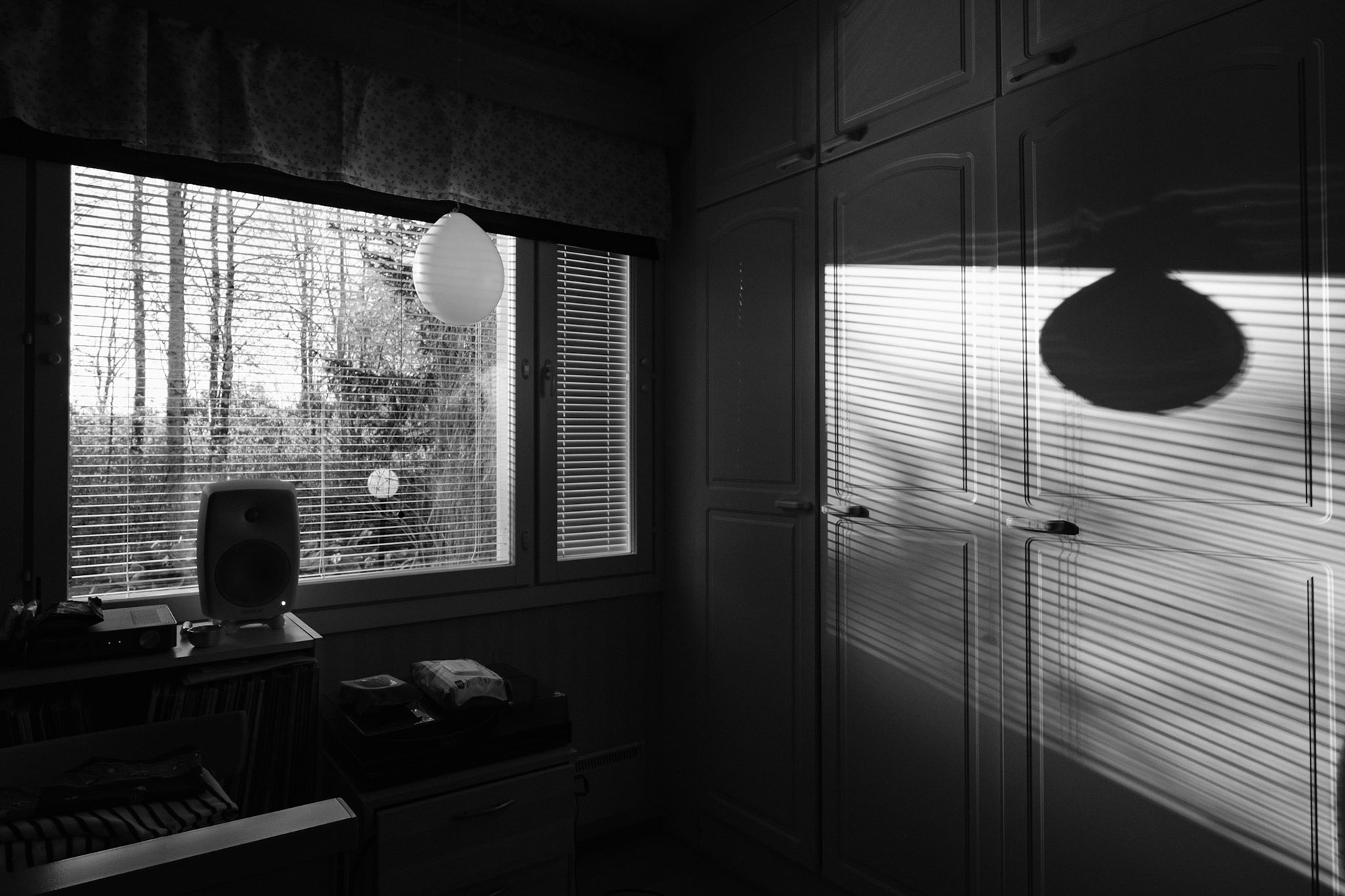 A black and white photo of a window view revealing a springy landscape. Inside the room, there is a balloon hanging in front of the window, which casts a long shadow onto a closet. There is also a turntable and a speaker, and various personal belongings on top of a sideboard.