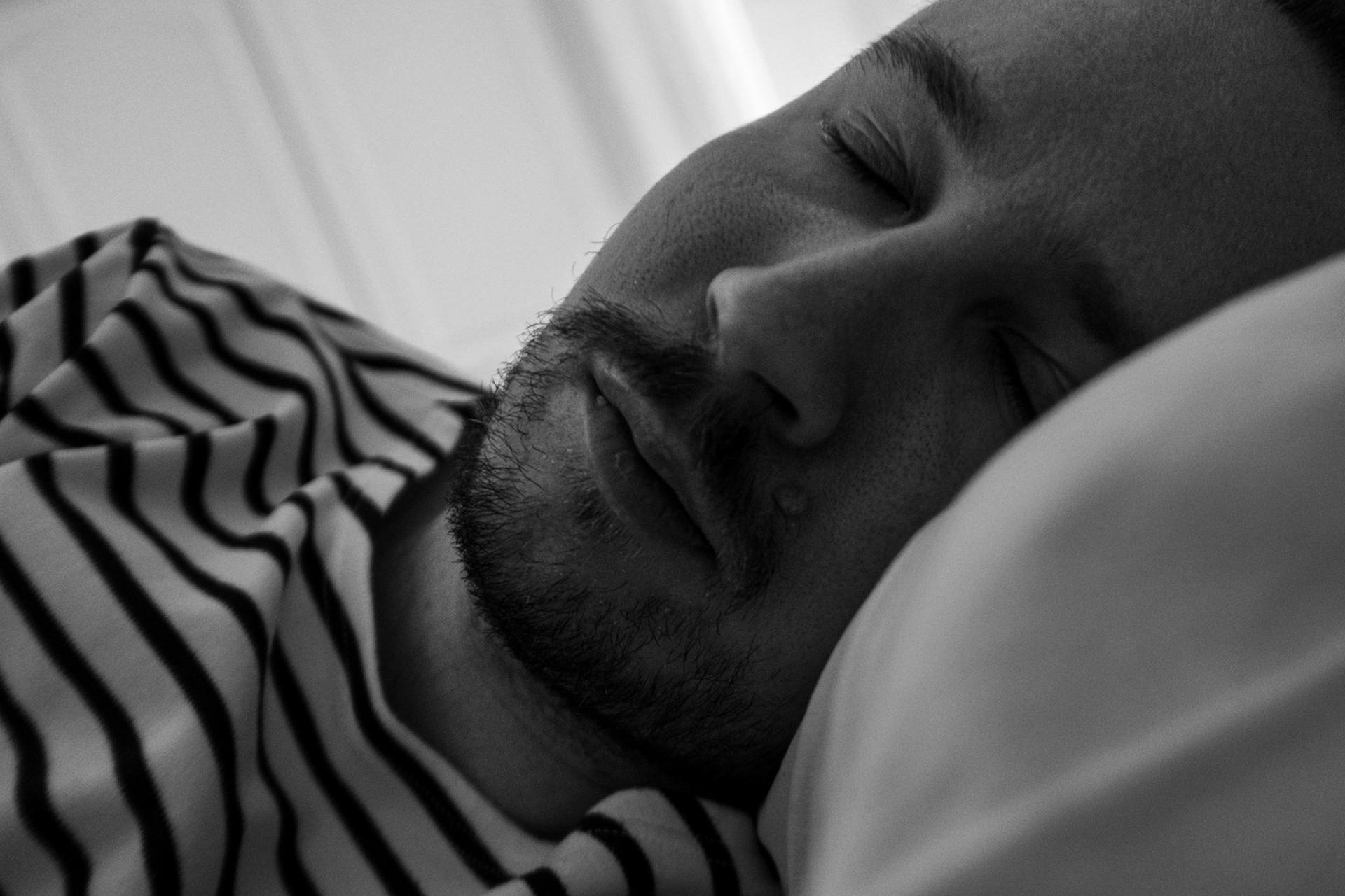 A black-and-white close-up photograph of Niko’s face while he rests his eyes closed in bed.