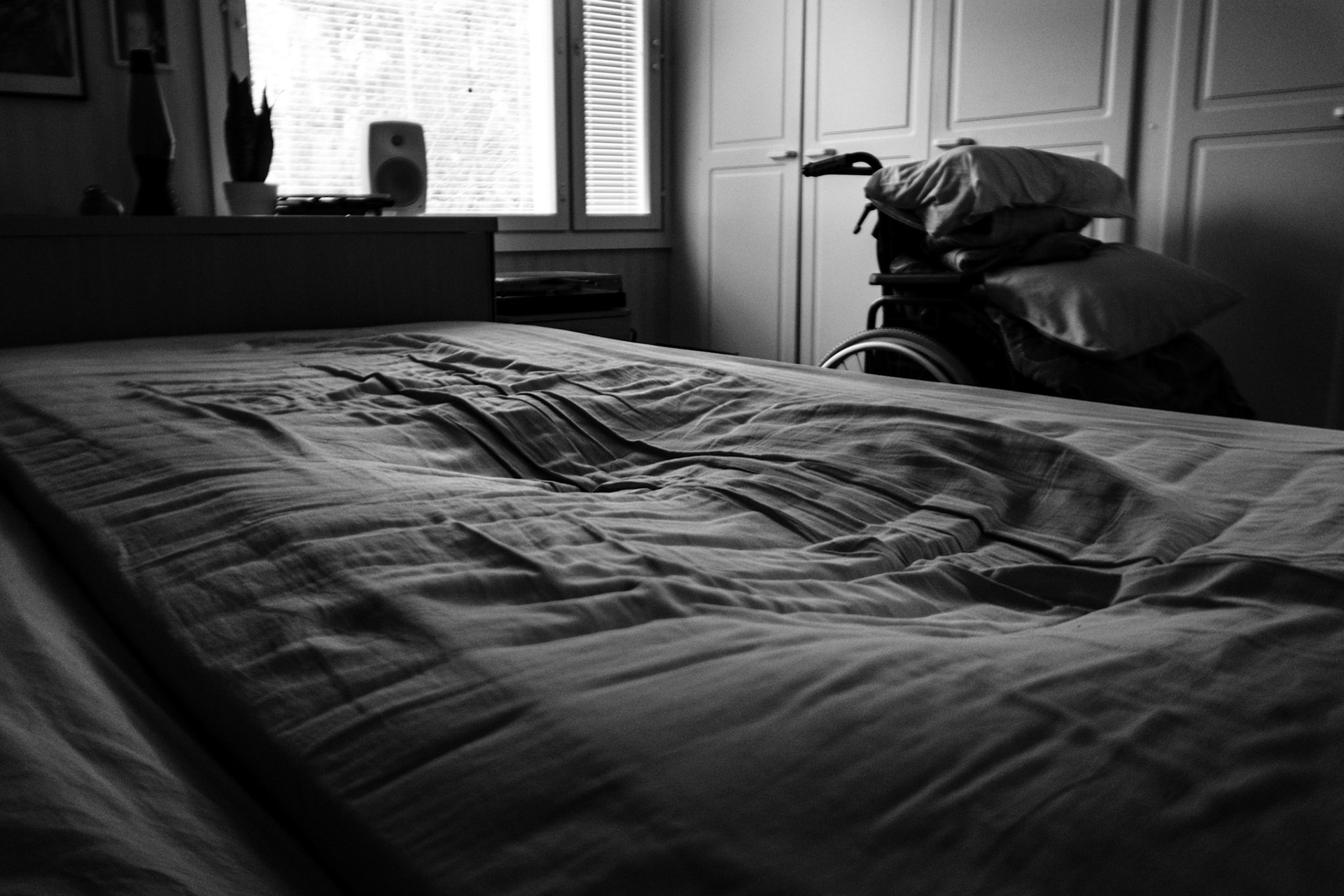 A black-and-white photo of an empty bed, which shows the dip of a human silhouette. A wheelchair, personal belongings, and a window are seen in the background.