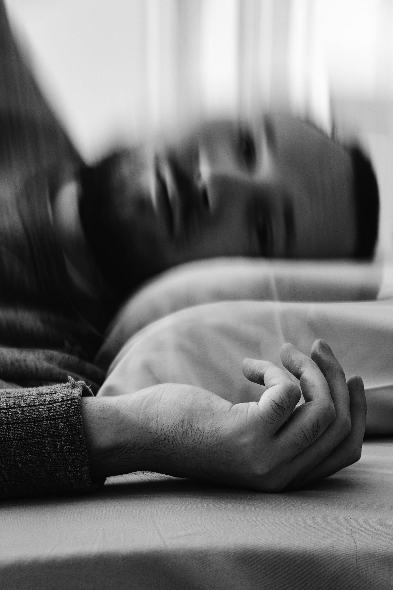 A black and white photograph of a man, Niko, resting his head on a pillow. His hand is in the foreground lying against the bed. His face appears blurry and distorted in the background.