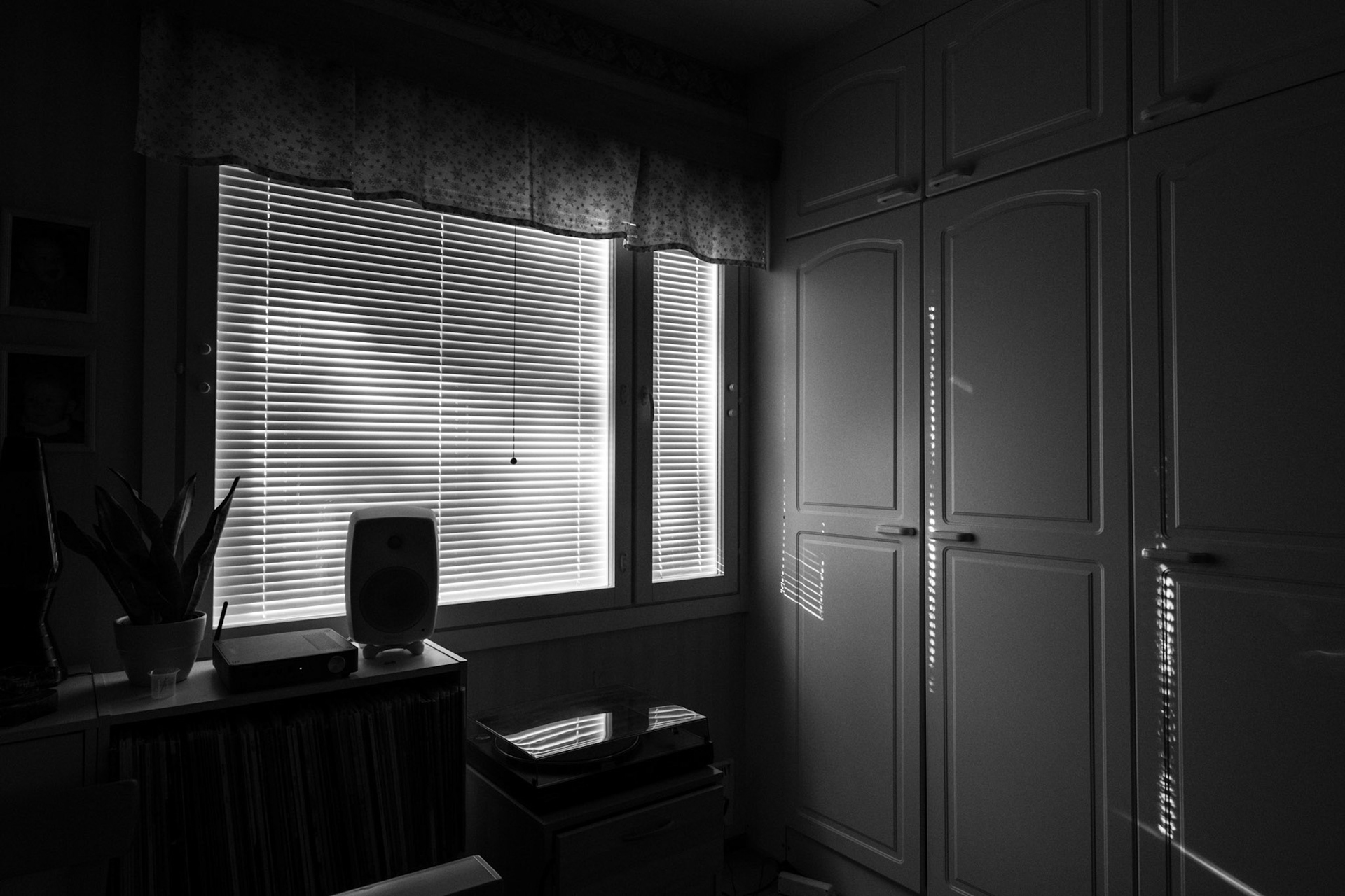 A black-and-white photograph of a dark bedroom corner: the blinds on the window are shut, and some abstract but small light patterns are visible on a closet next to the window.