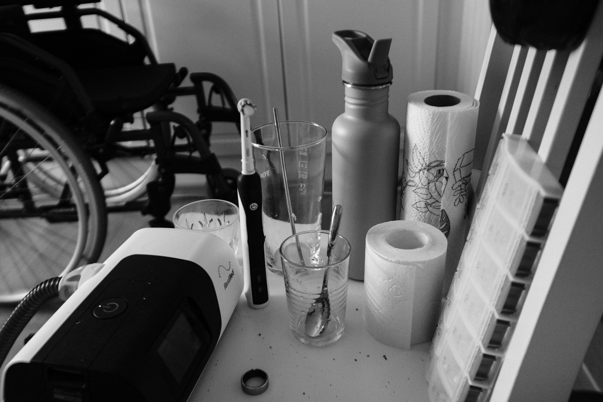 A black and white photo of a cluttered chair with an electric toothbrush, water bottle, paper towel roll, pill organizer, and other personal items. A wheelchair is in the background.