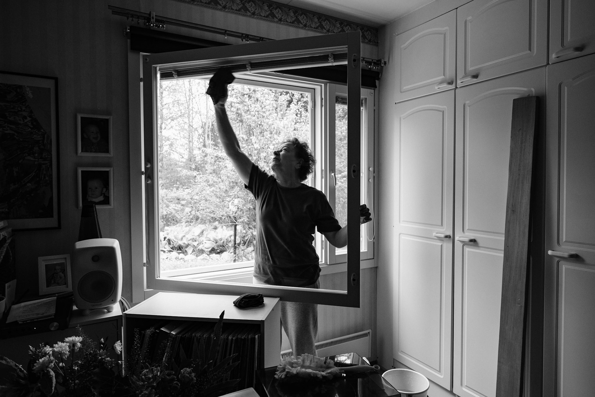 A black and white photo of an older woman washing an open window inside a bedroom. She’s facing the camera and reaching with her hand to wash the upper part of the window. Light comes through the open window. Personal belongings are visible on a shelf.
