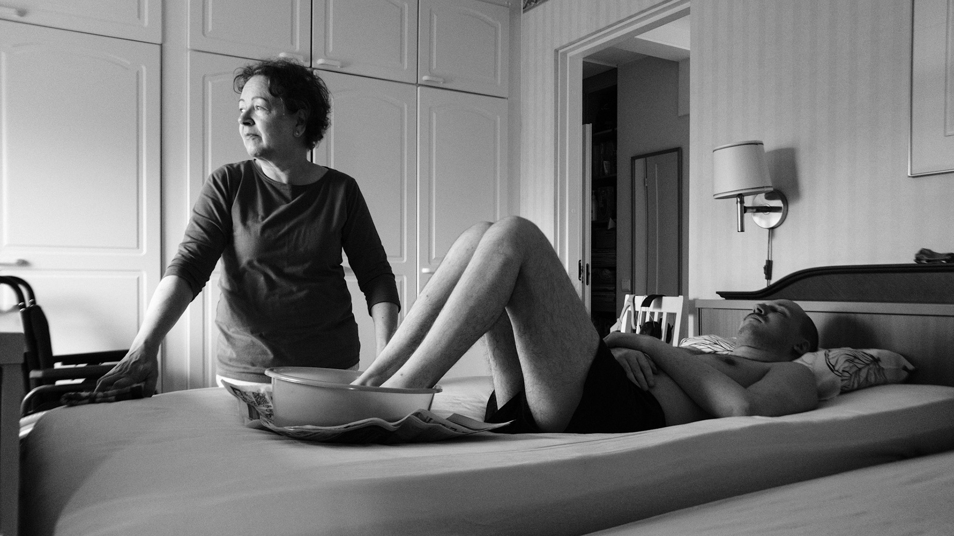 A black and white photo of a woman, Päivi, looking outside of the frame as she prepares to wash a man’s, Niko’s, feet as he lies on a bed his feet in a basin wearing just boxers. The room features white cabinets and is a bedroom.