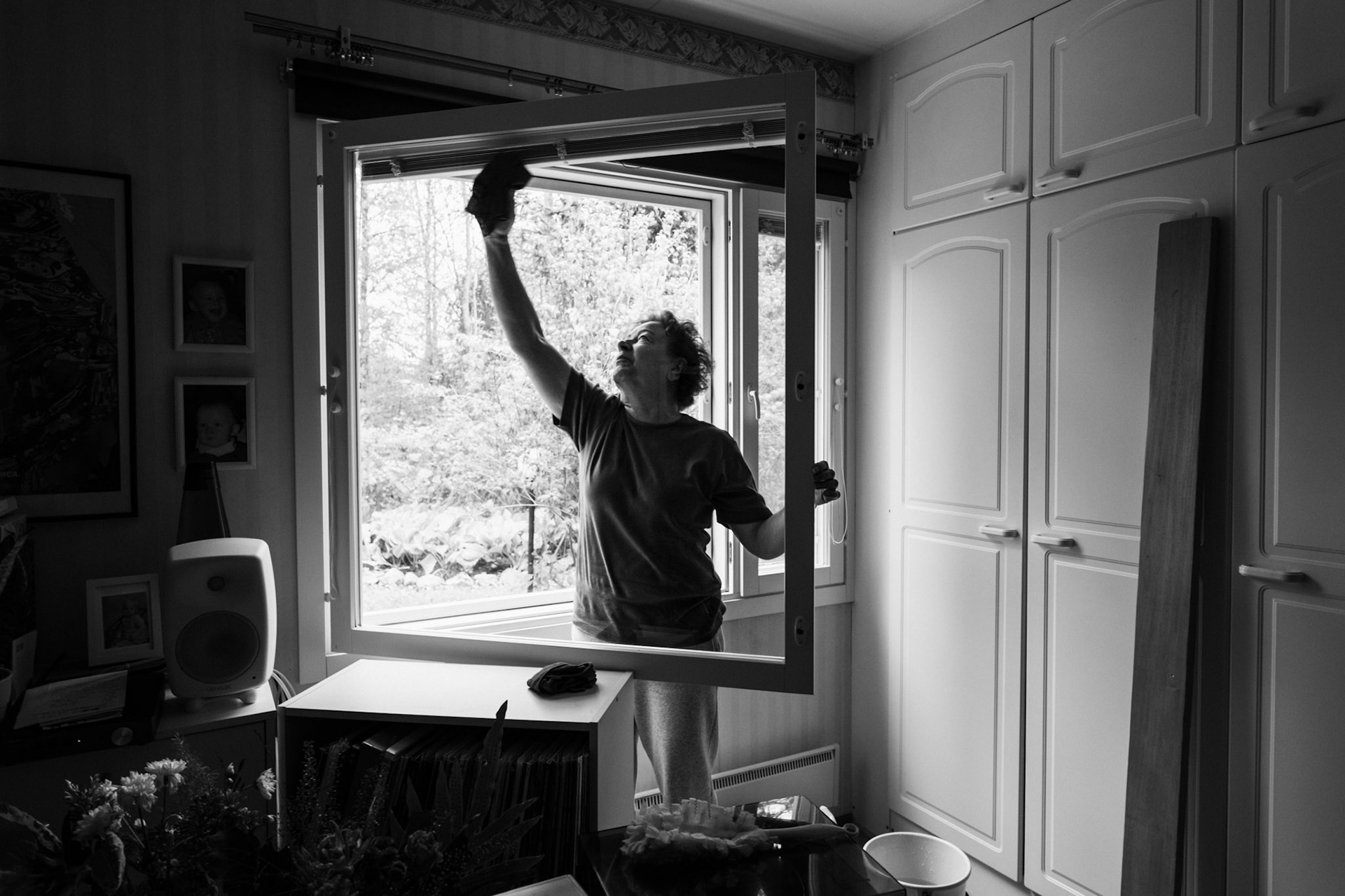 A black-and-white photo of an older woman washing an open window inside a bedroom. She’s facing the camera and reaching with her hand to wash the upper part of the window. Light comes through the open window. Personal belongings are visible on a shelf.