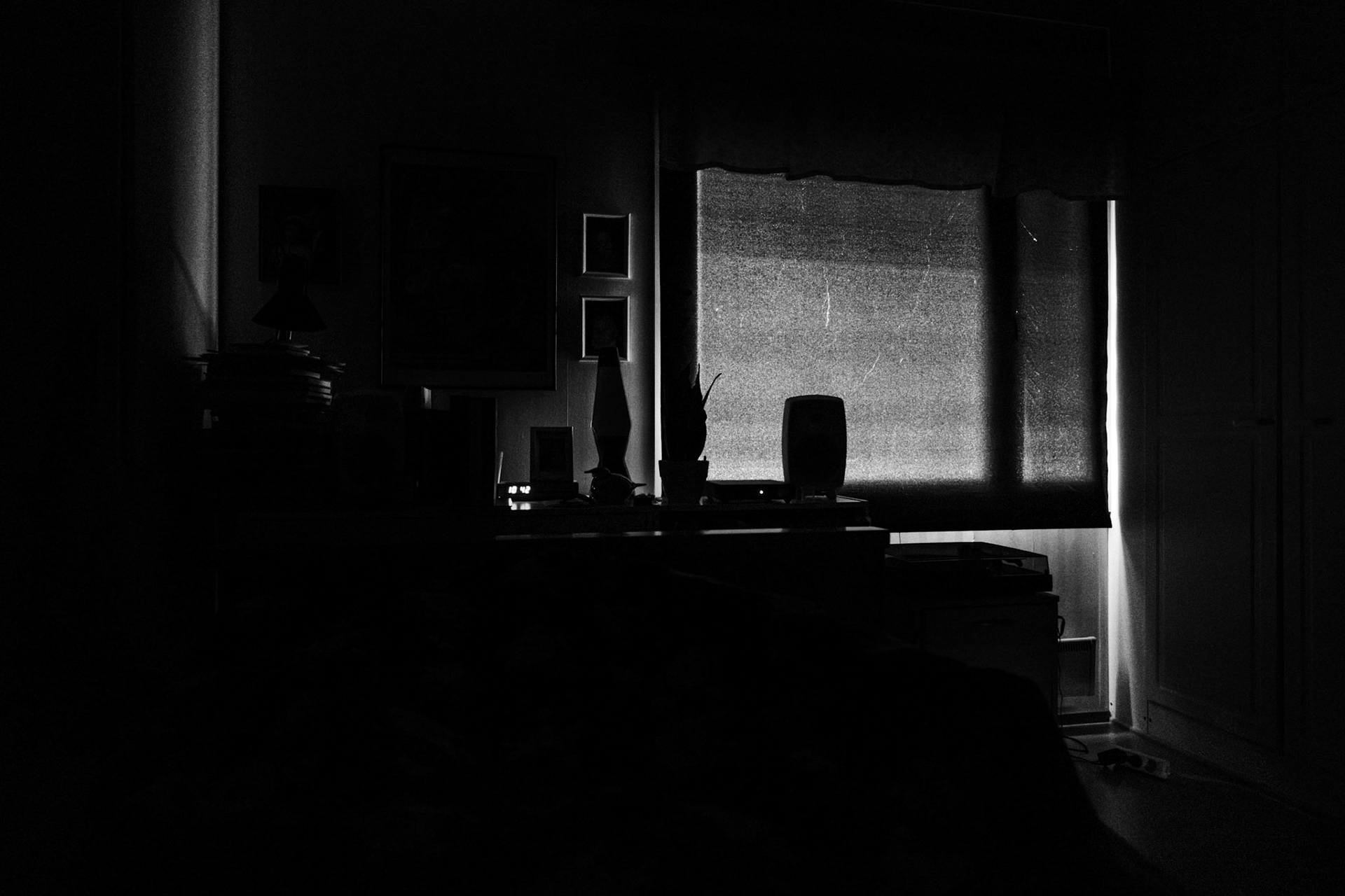 A high contrast black-and-white photograph of a completely dark bedroom. A long and narrow ray of light peaks through the corner of the curtain-covered window. The silhouette of the window is seen through the curtain.