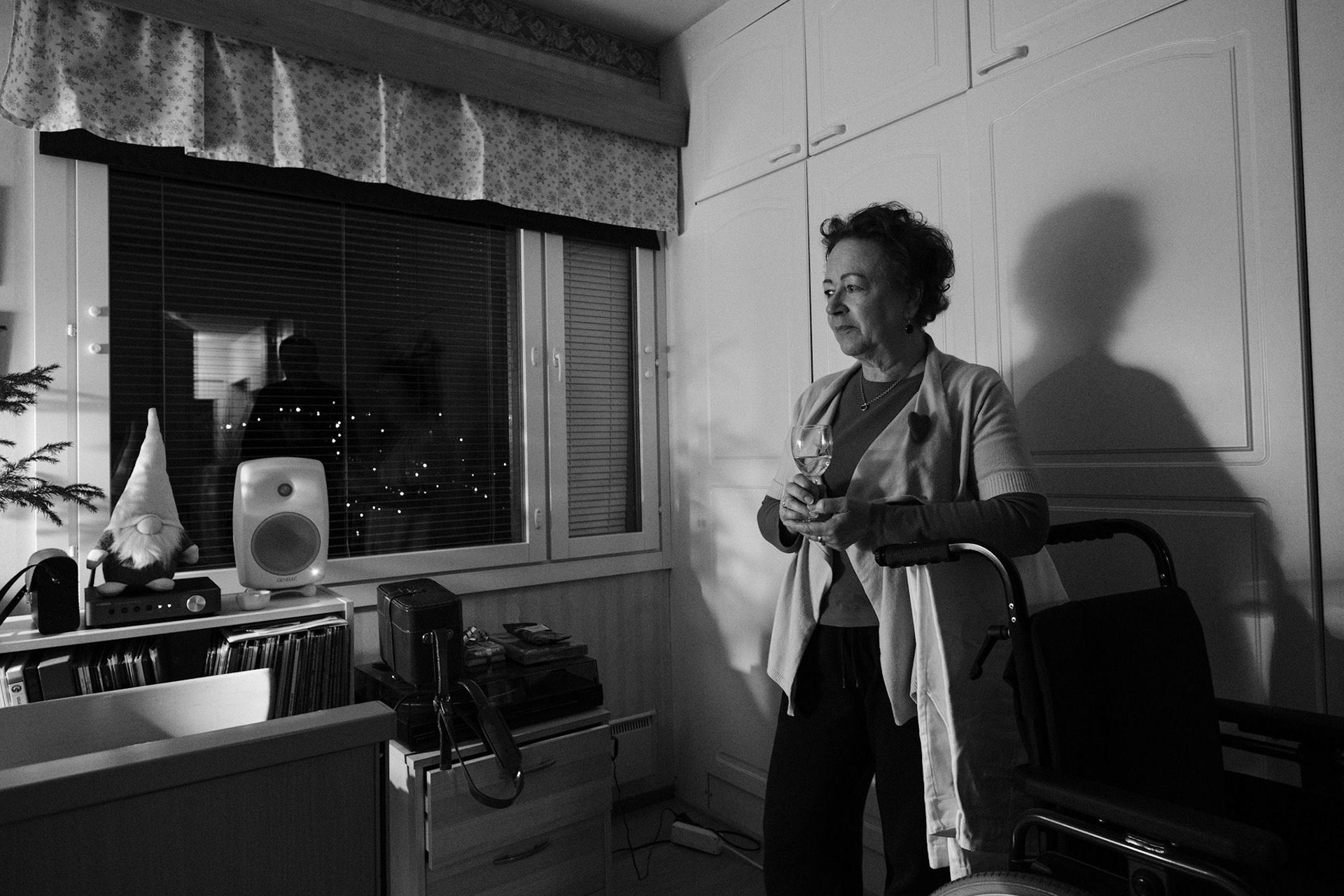 A black and white photo of Päivi, a woman, with short, curly hair, standing in a dimly lit room holding a wine glass. She leans on a wheelchair. Her shadow falls on white cabinets. A window with blinds reflects another silhouette.
