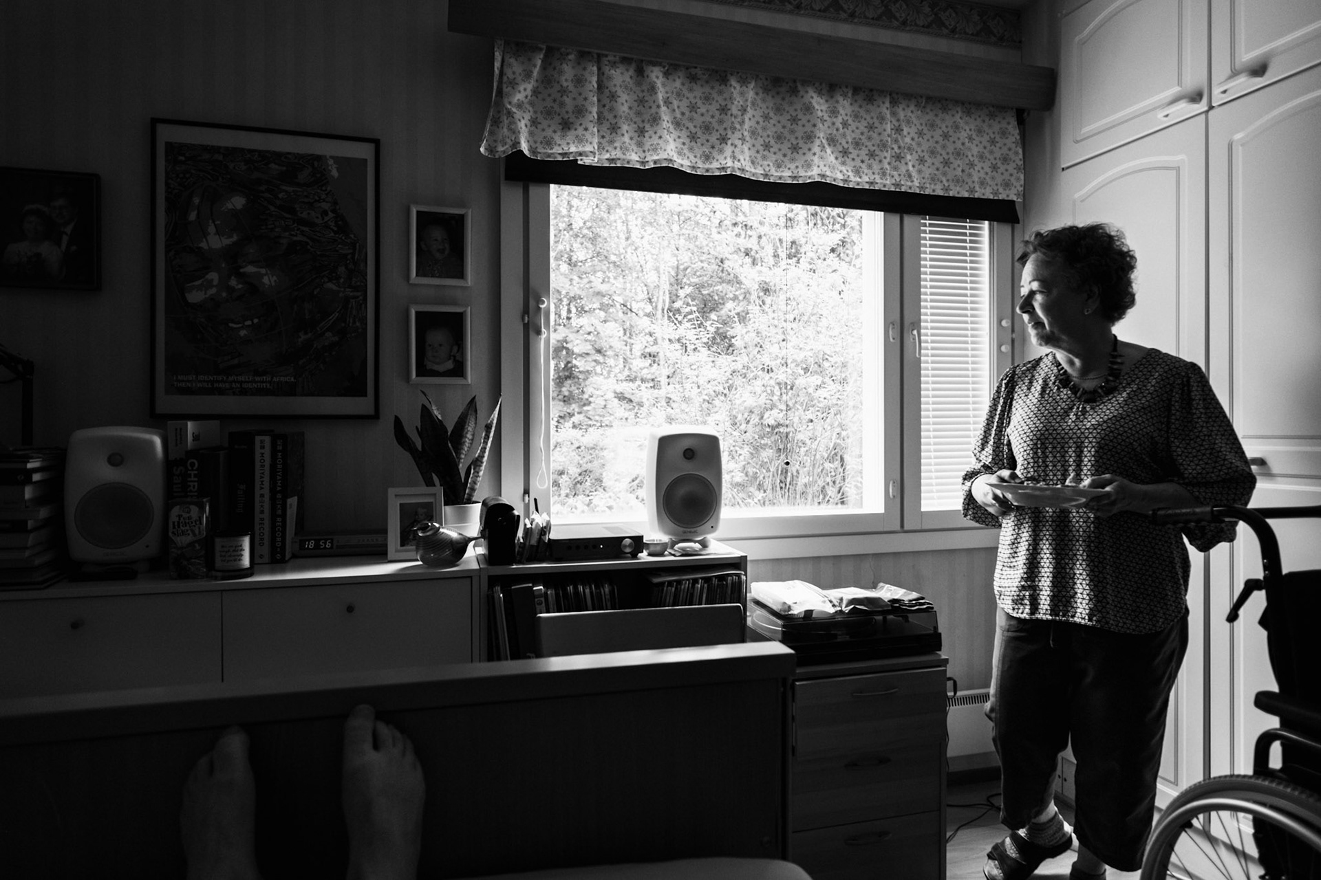 A black-and-white photo of a window view revealing a summer landscape. Inside the room, Niko’s mother has brought him his lunch. She is holding a plate in her hands. There is a turntable and a speaker, and various personal belongings on top of a sideboard.